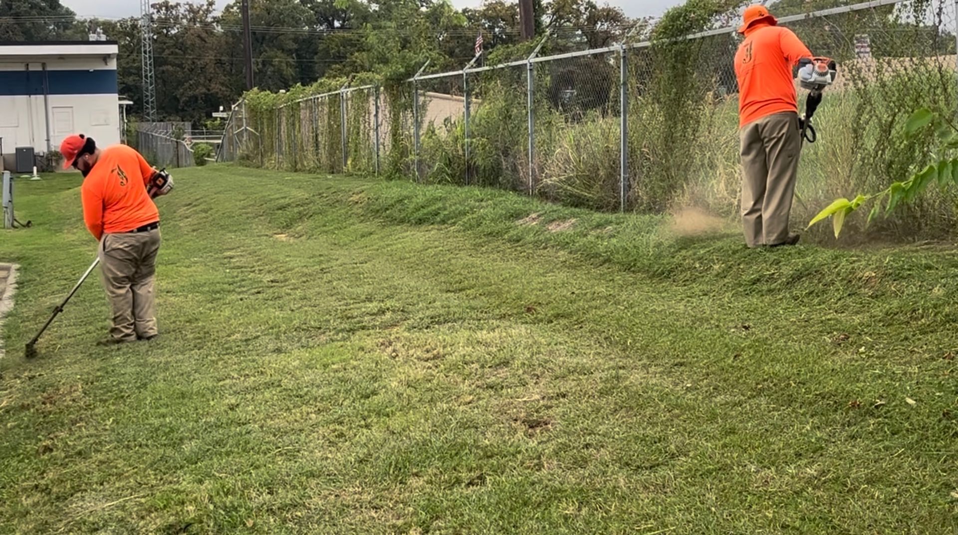 Two workers in orange suits using weed eaters to trim grass near a chain-link fence.