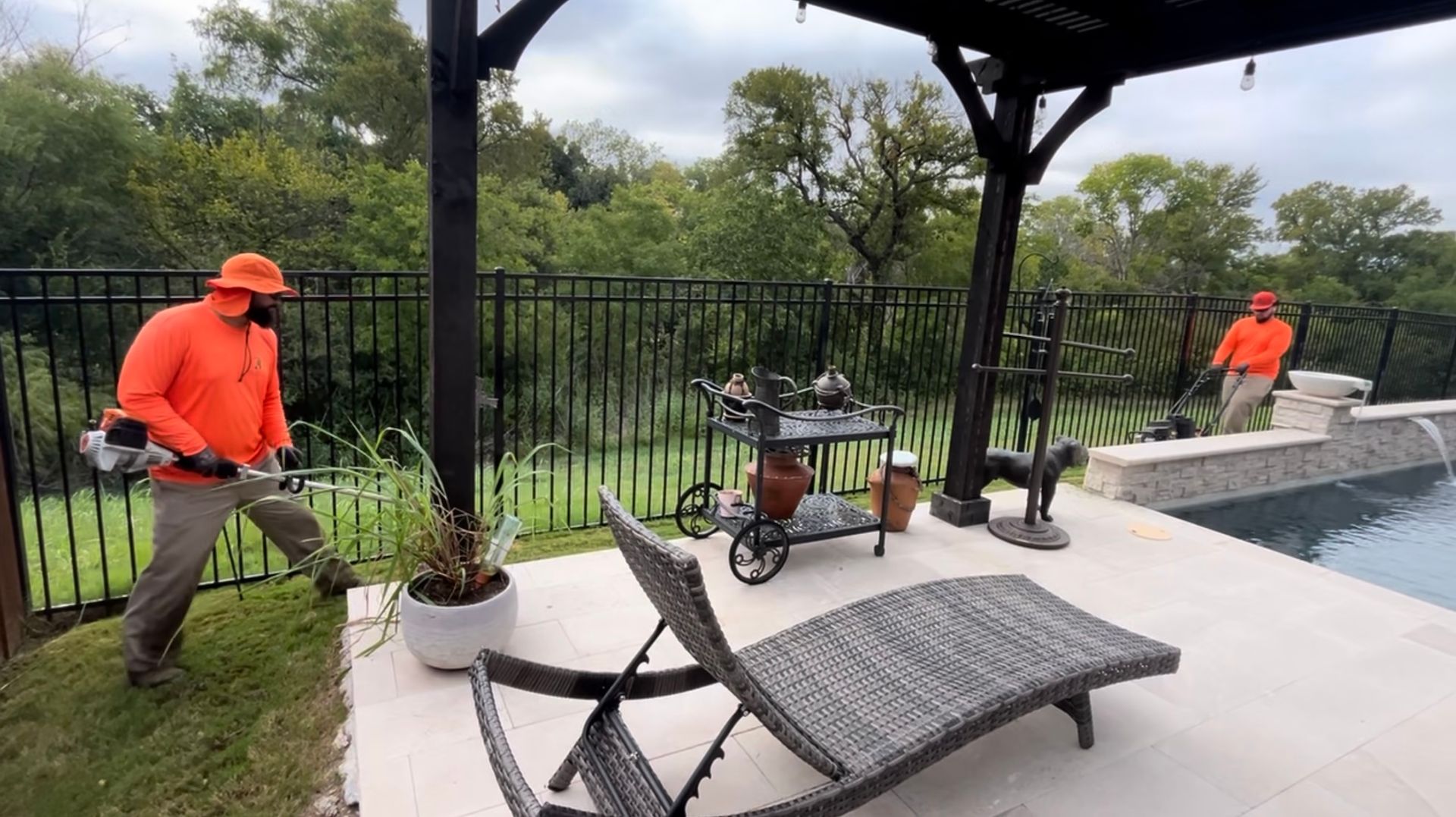 Two people in orange shirts trim grass near a pool, black fence, and pergola.