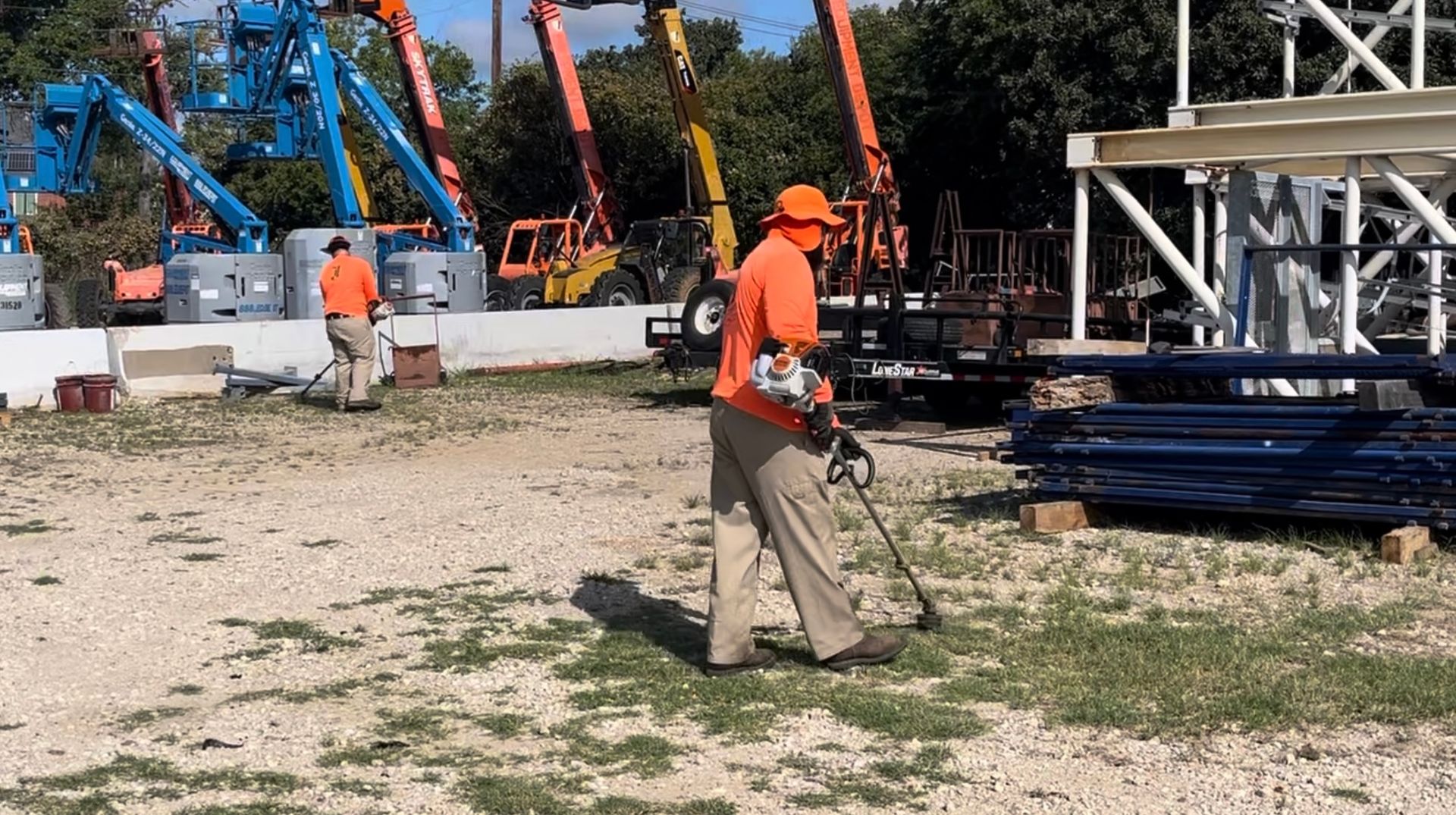 Two workers in orange shirts use weed trimmers on a gravel lot with equipment in the background.