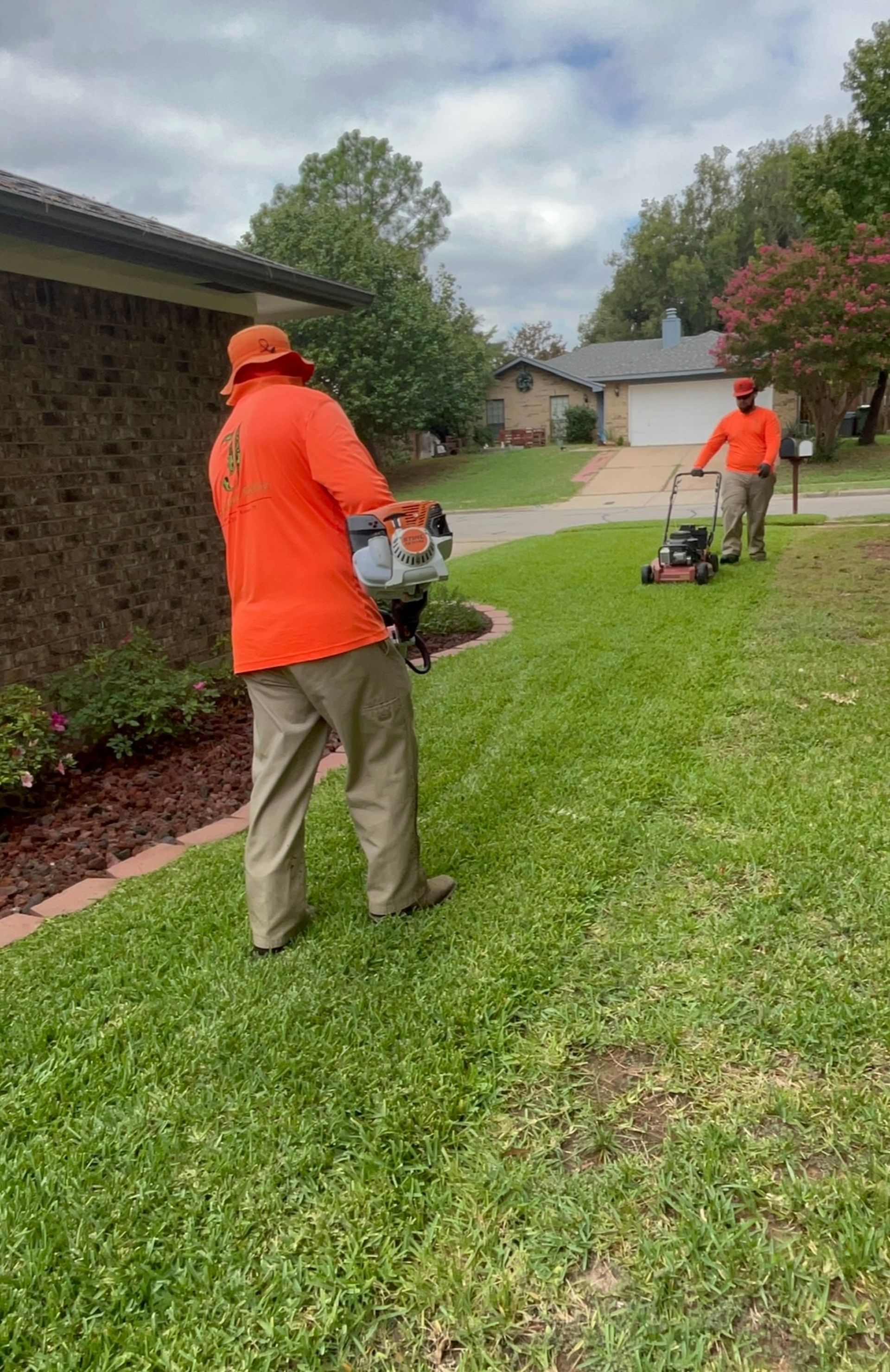 Two men in orange shirts mowing a lawn; one uses a weed eater, the other a mower.