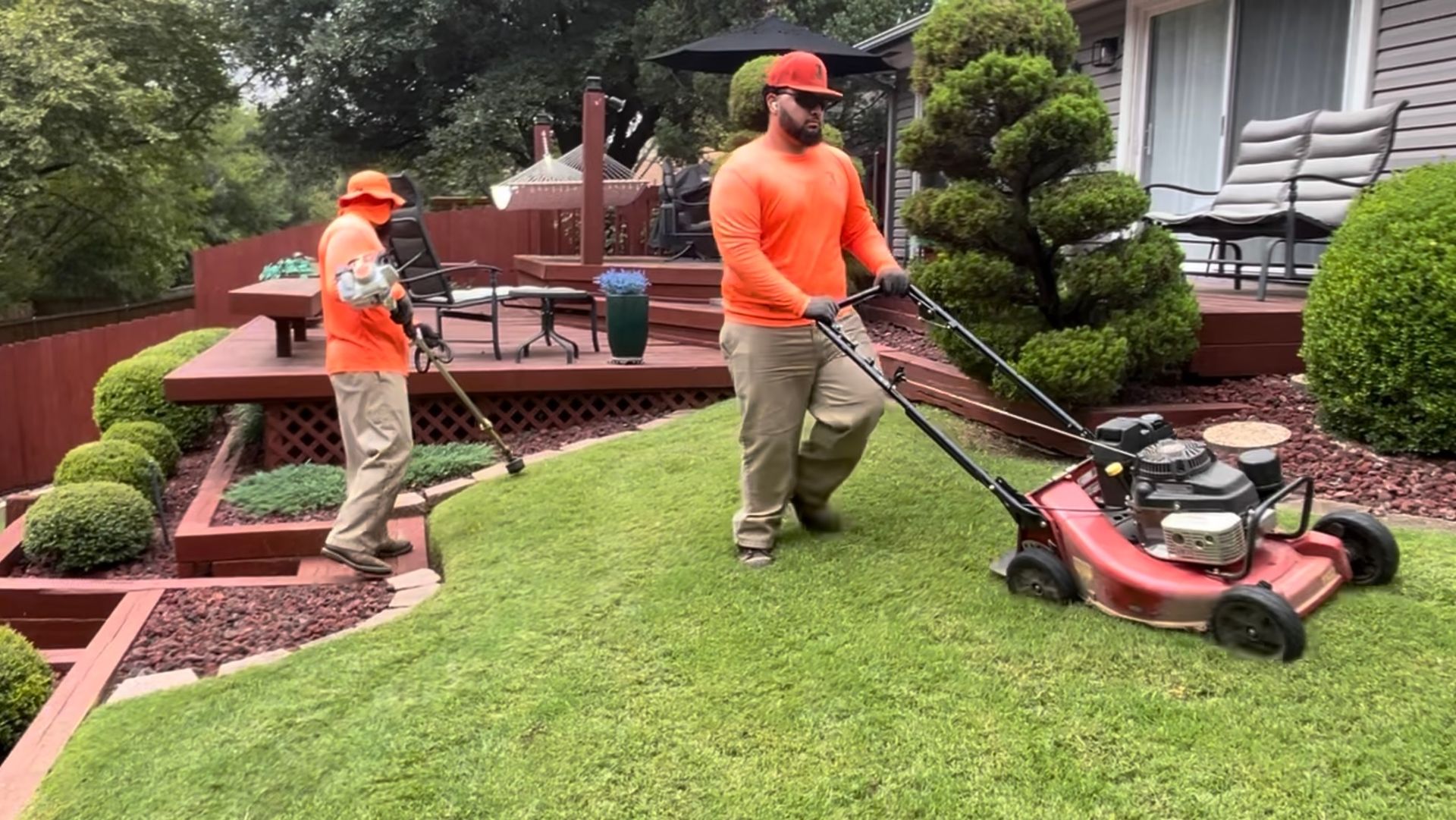 Two landscapers mowing a lawn: one with a mower, one with an edger. Both wear orange shirts and hats. Green grass.