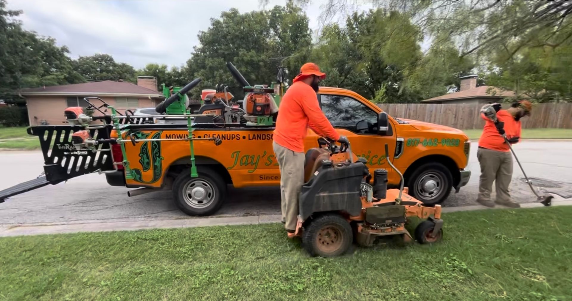 Two landscapers in orange shirts and hats mowing grass beside an orange work truck.