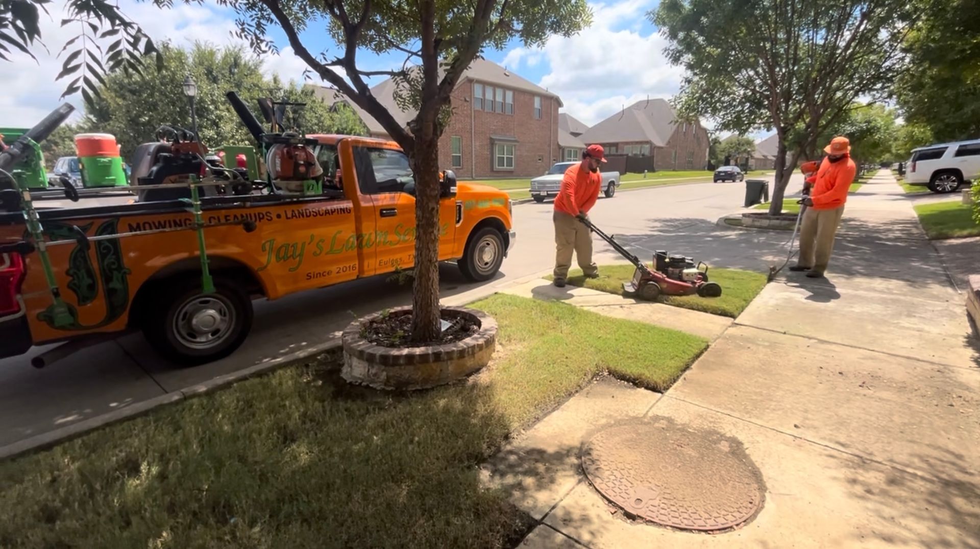 Two landscapers lay sod on a sidewalk next to an orange truck with equipment in front of a suburban house.