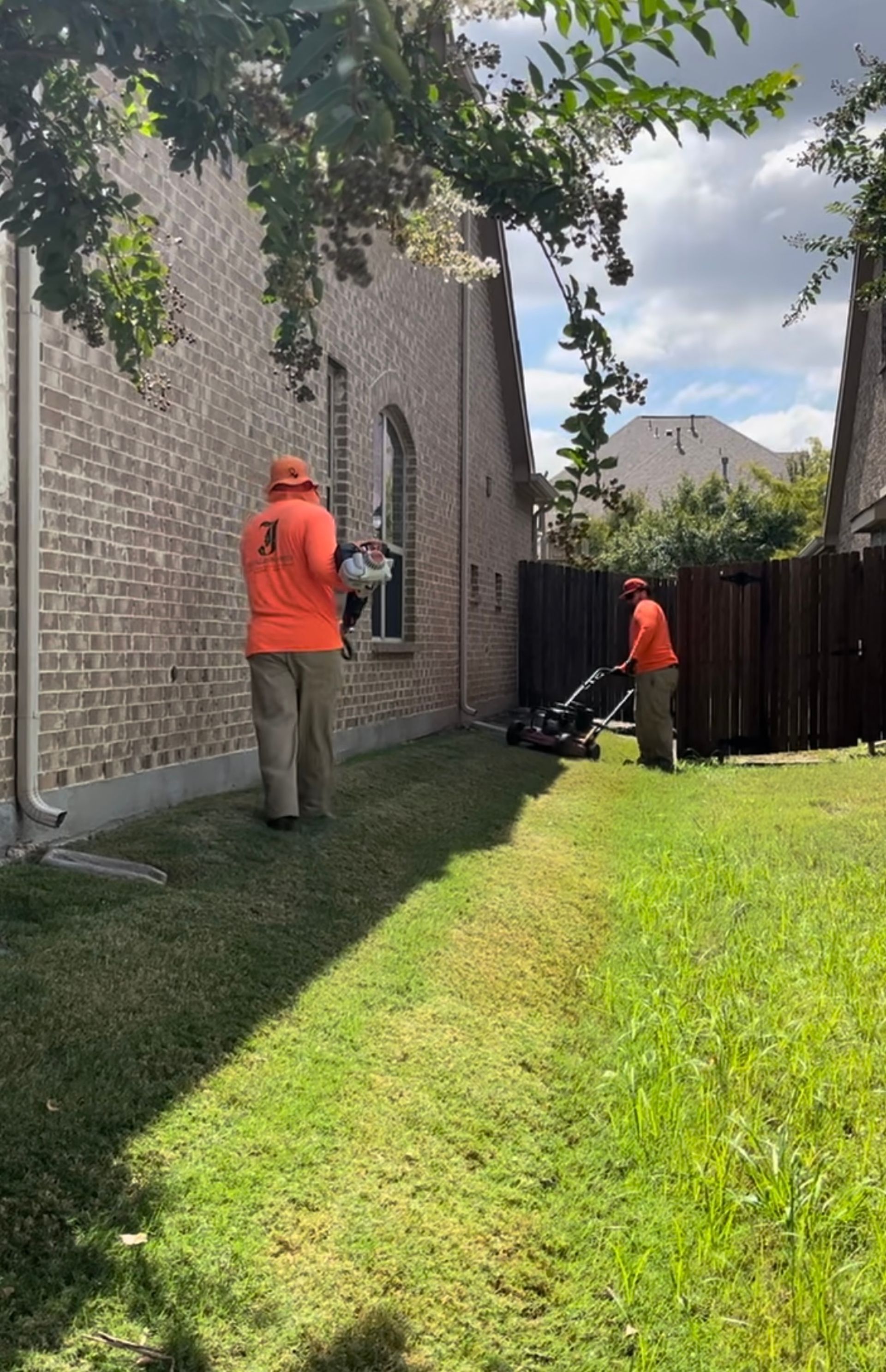 Two people in orange shirts mowing a lawn next to a brick house on a sunny day.