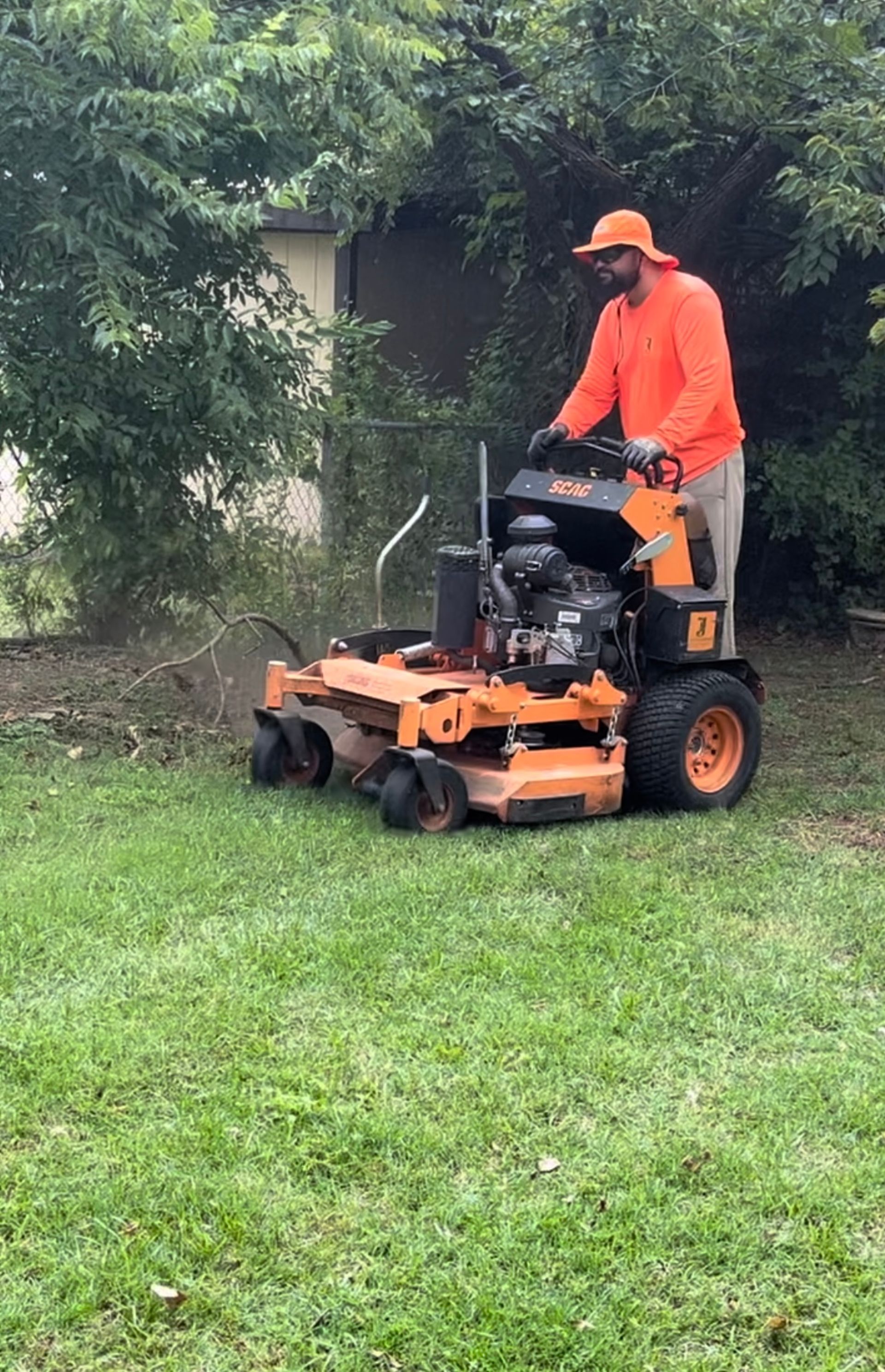 Man in orange mowing lawn with an orange lawnmower near a chain-link fence and greenery.