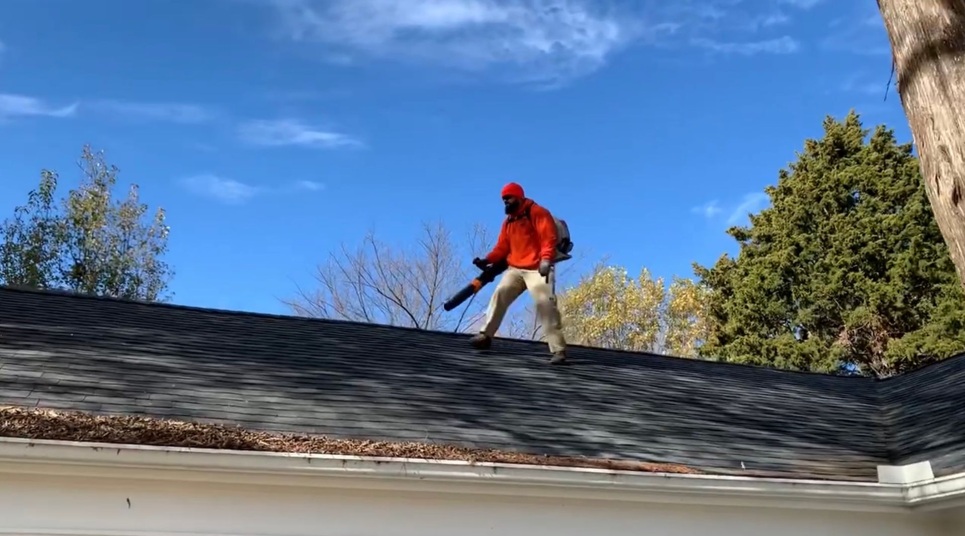 Person in orange jacket uses leaf blower on a rooftop, blue sky above.