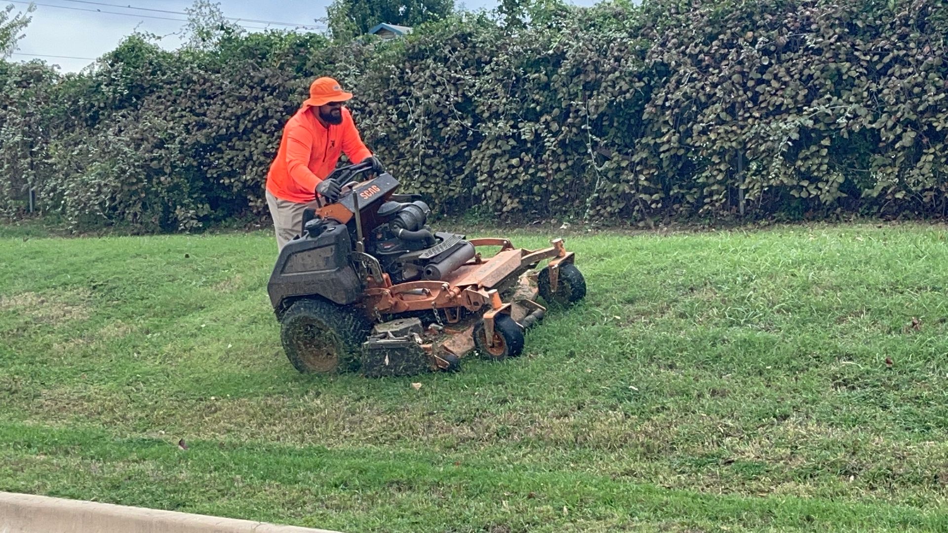 Man in orange shirt mows grass with riding mower on a grassy area near bushes.