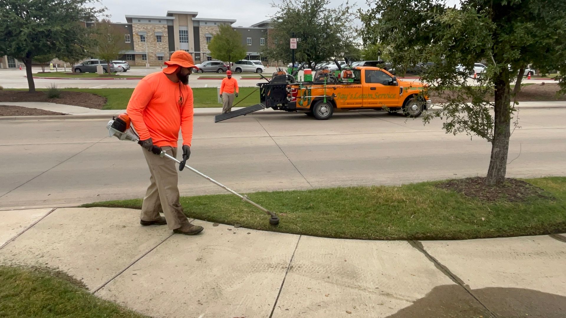 A worker in orange uses a weed wacker on grass near a sidewalk and road. Other workers and a truck are visible.