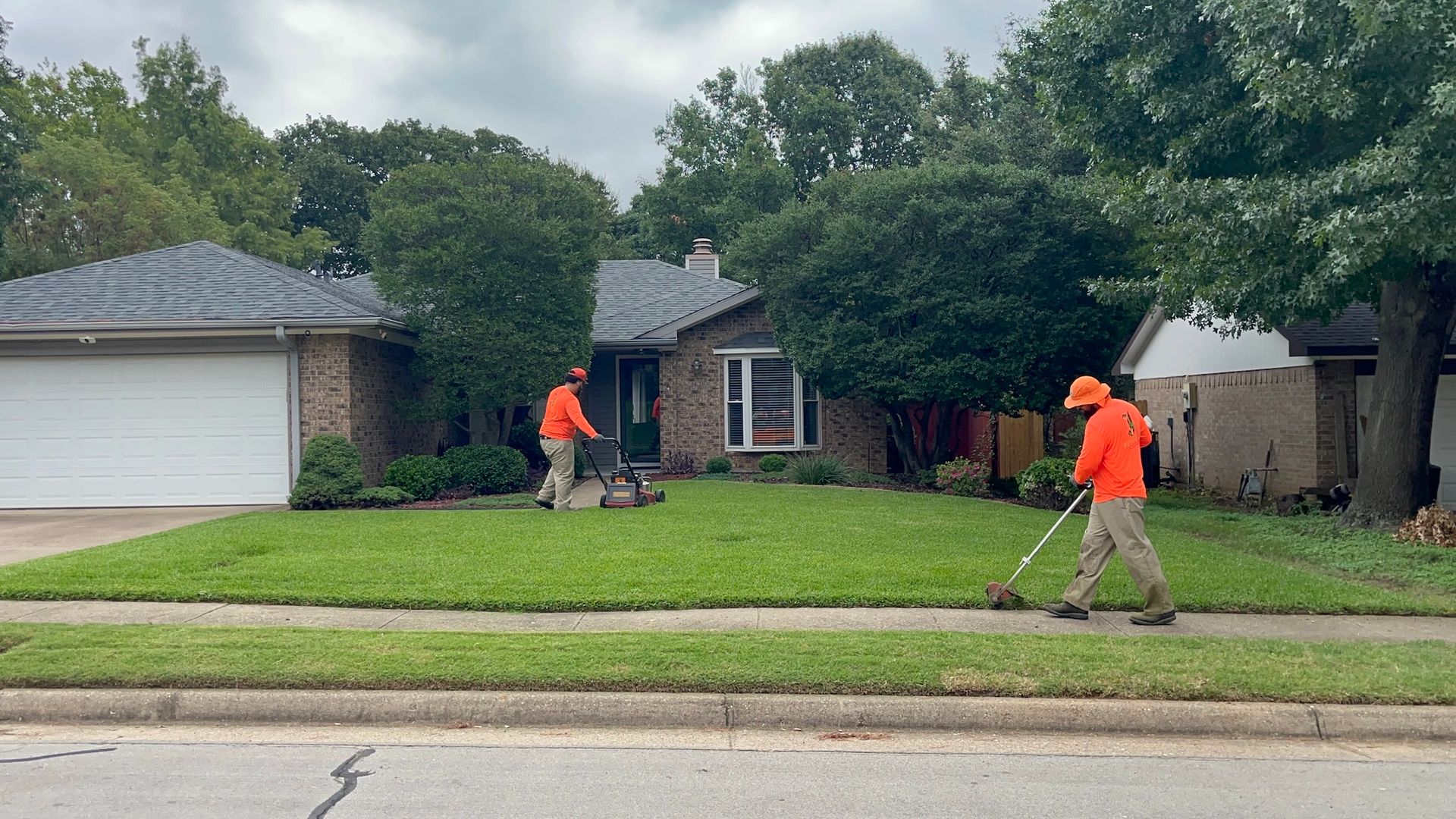 Two landscapers mowing a lawn in front of a house, one using a mower, the other a string trimmer.