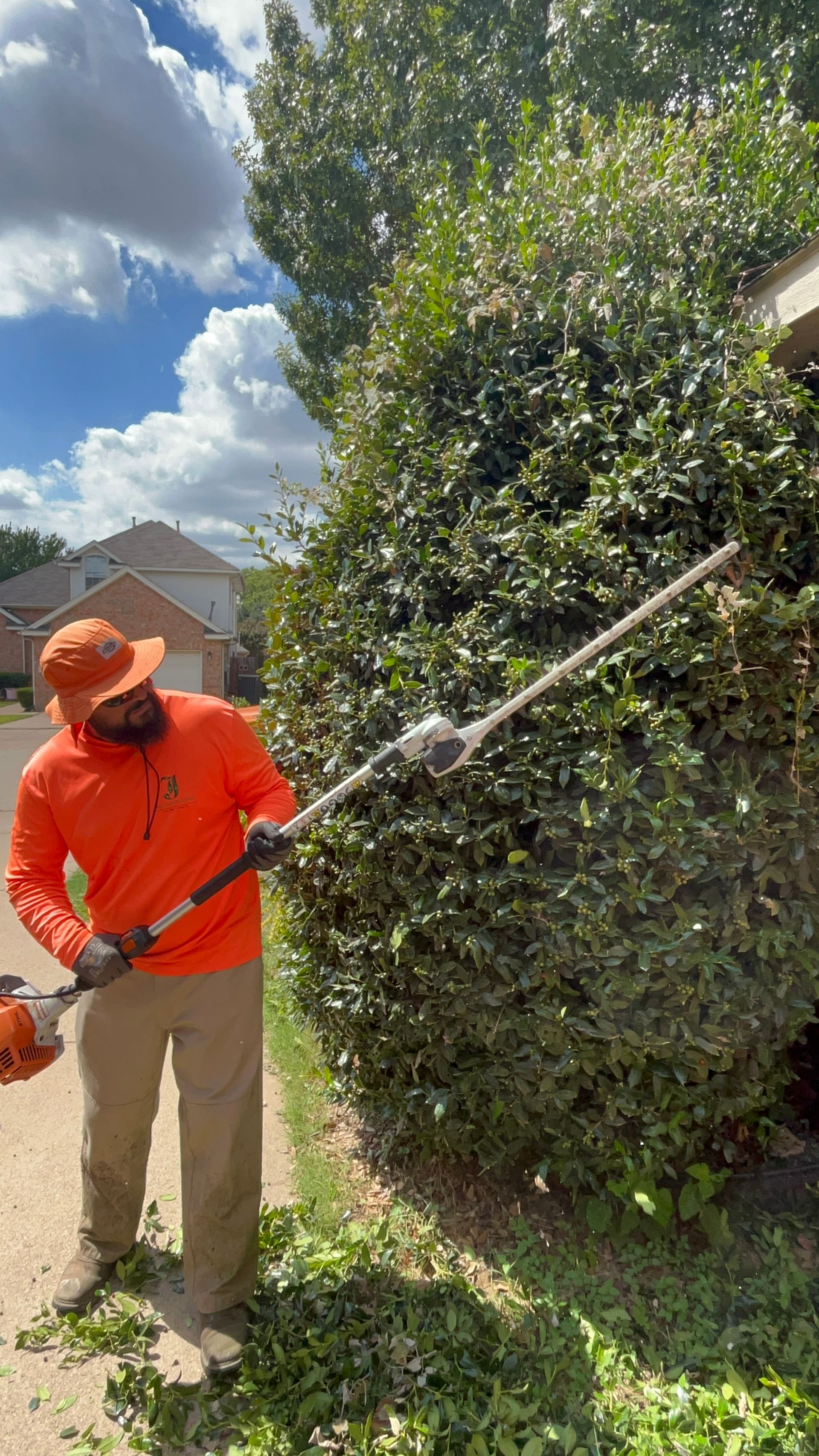 Man in orange trimming a tall green bush with a pole hedge trimmer on a sunny day.
