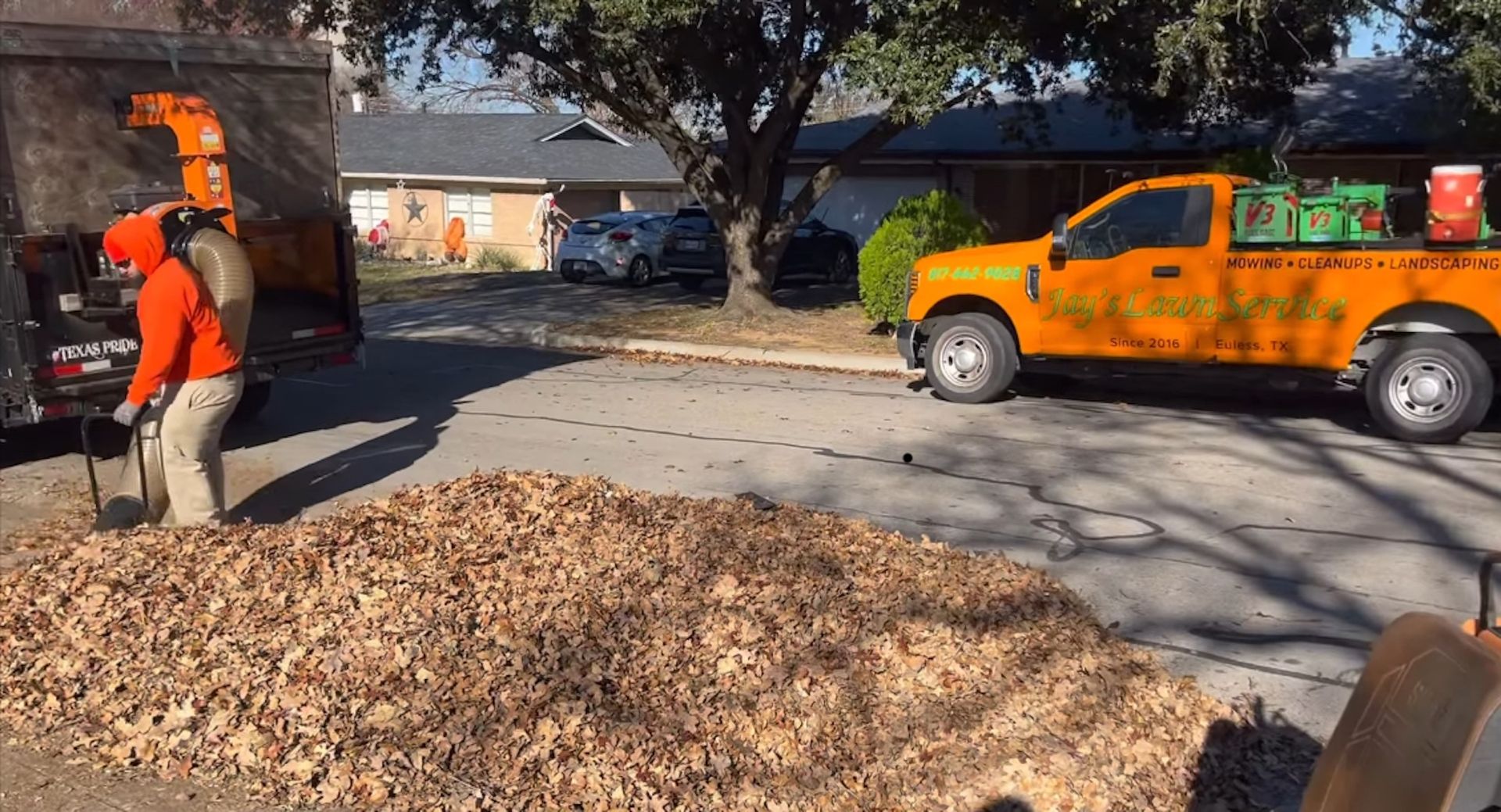 Man using a leaf blower to move leaves into a truck on a residential street. Orange truck, brown leaves.