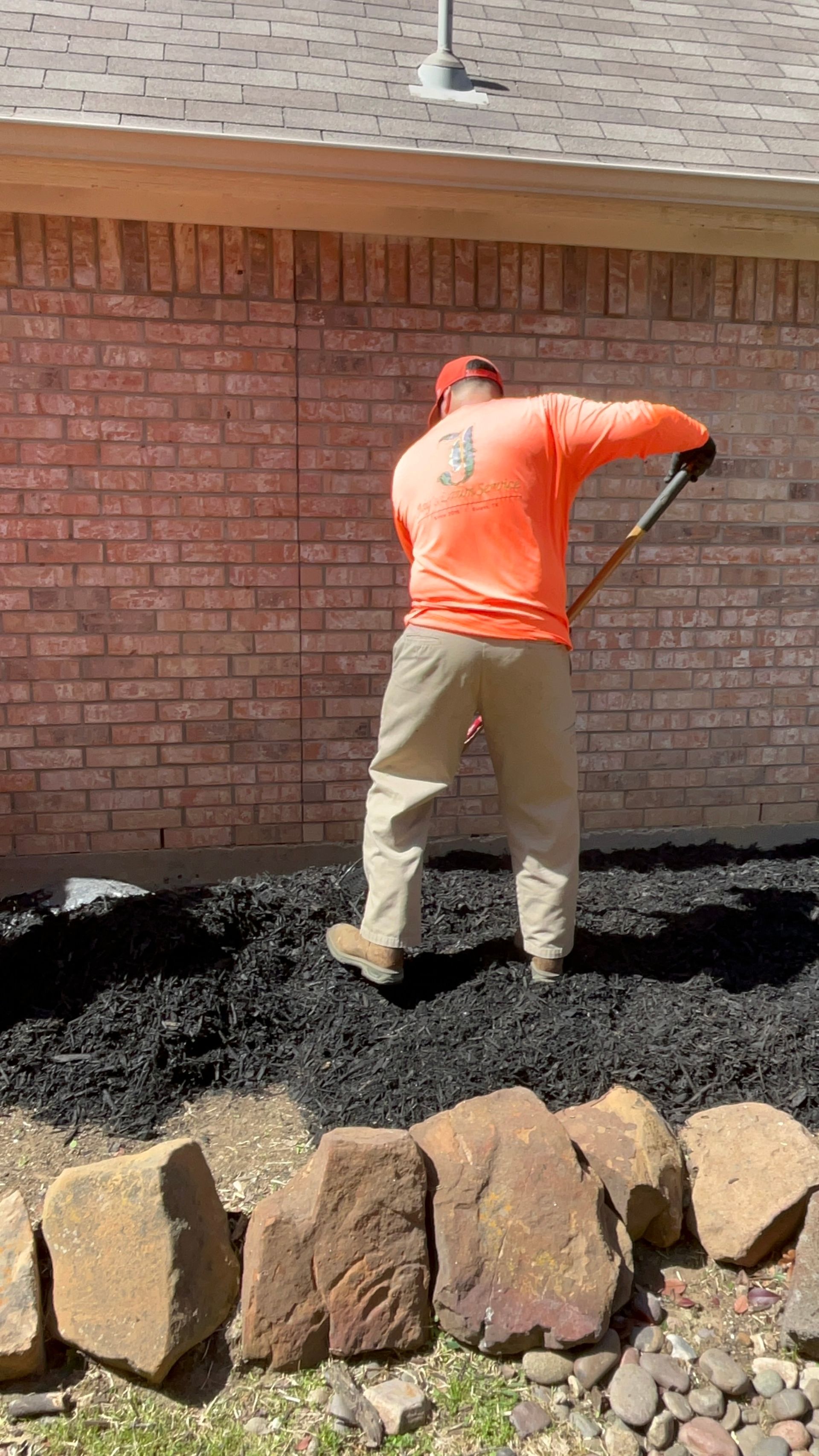 Man in orange shirt spreading black mulch against a brick wall using a long handled tool.