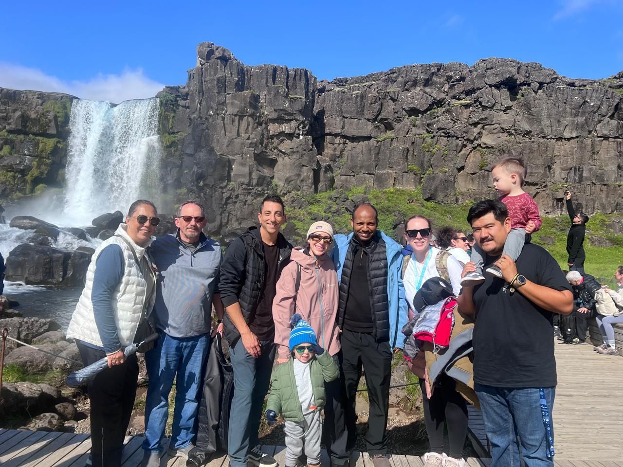 A group photo showing tourists from Arizona posing on a wooden walkway at the base of Öxarárfoss waterfall in Þingvellir (Thingvellir) National Park, Iceland. The waterfall cascades over the black basalt rocks of the Almannagjá gorge under a bright blue sky. The diverse group, including a toddler on an adult's shoulders and a young child in front, are on a Celebrity Silhouette cruise guided Golden Circle shore excursion.