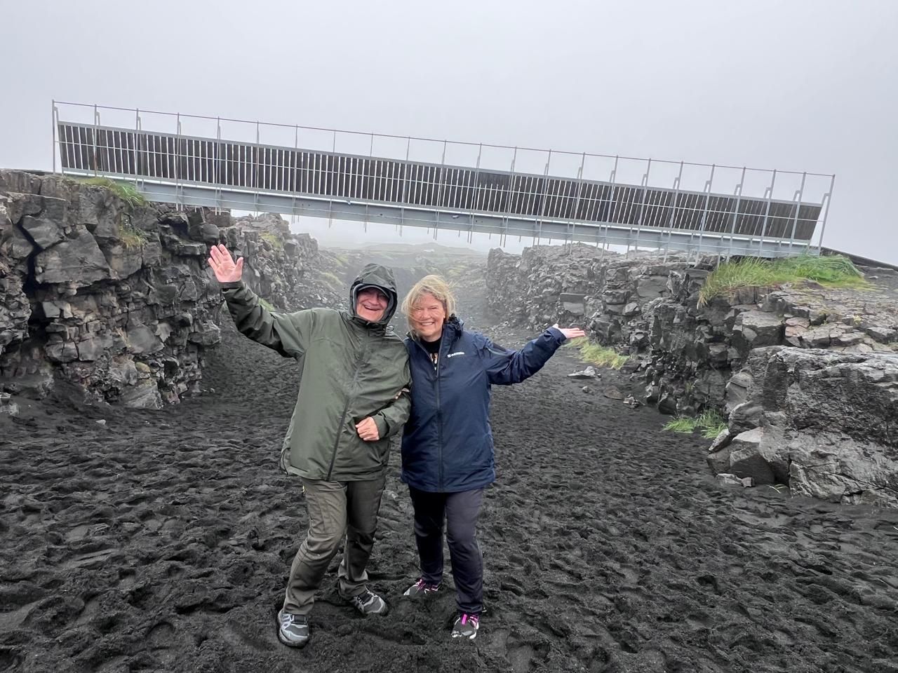 A photo  showing two smiling tourists, including a waving woman, from Colorado, USA. They are dressed in hooded hiking jackets and are on a guided tour in Iceland, posing at a wooden platform with a metal railing. In the background is a large, high metal and glass structure, possibly a bridge or weather station, positioned over a dark, rocky volcanic rift under a dense, foggy sky.