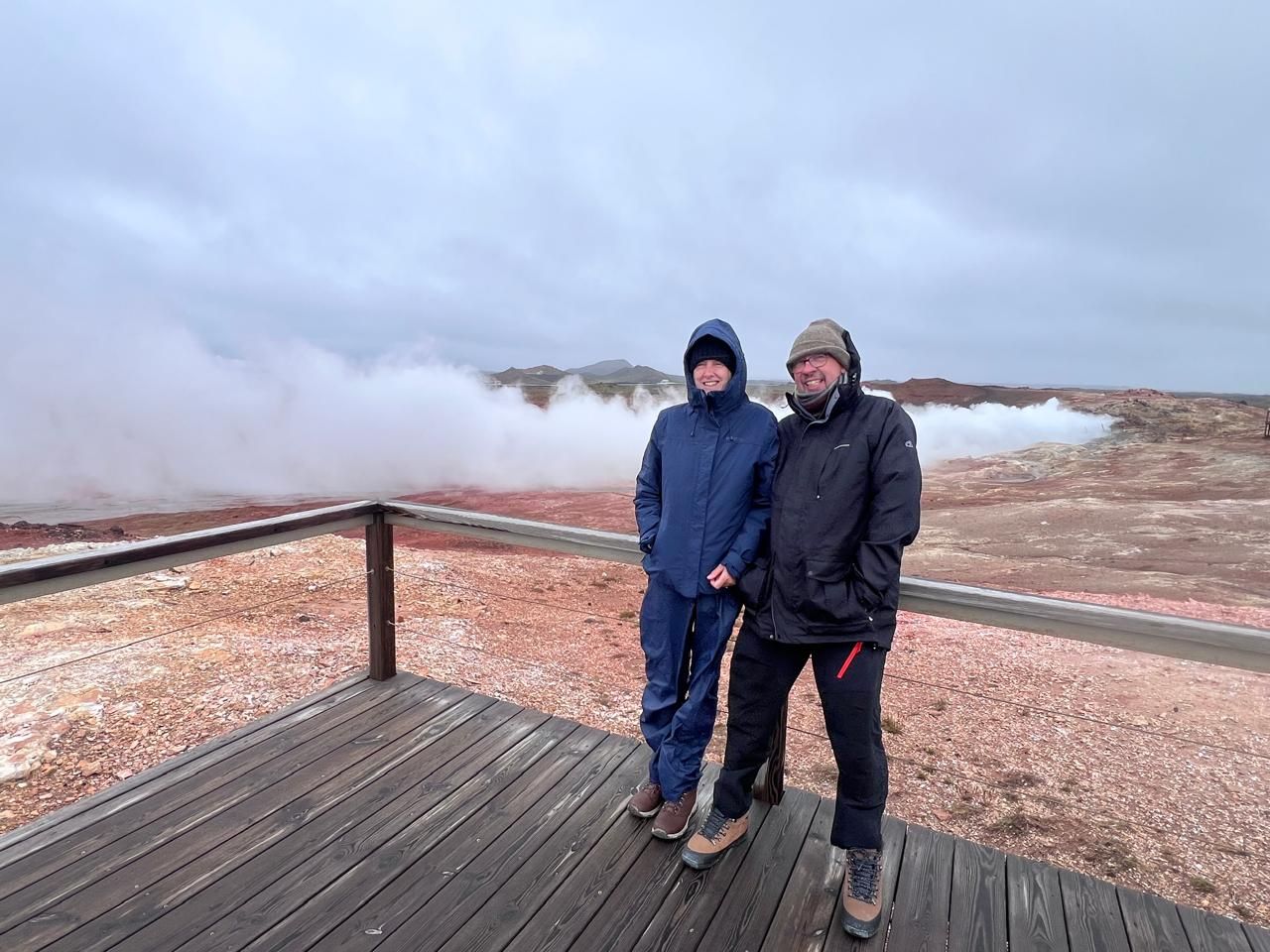 A smiling couple dressed in weather-appropriate hiking gear on a wooden boardwalk. They are posing at a viewing platform overlooking a dramatic geothermal field in the Reykjanes Peninsula, Iceland. The red, oxidized earth of the Gunnuhver area is visible, with heavy, white steam clouds rising against a grey, overcast sky. This image captures tourists from the Sky Princess cruise from England on a guided tour