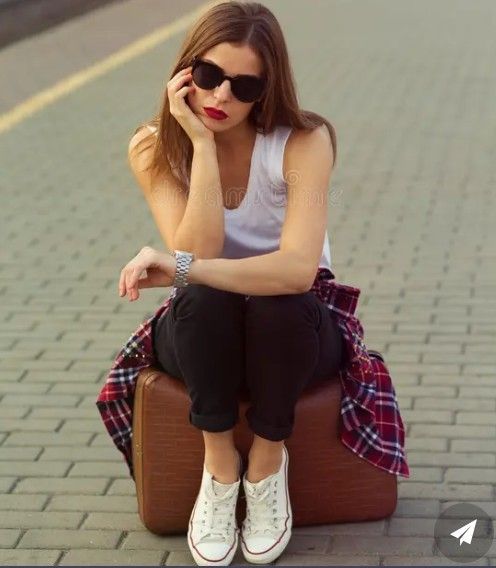 A woman wearing sunglasses, a white top, and black pants sits on a brown suitcase outdoors, checking