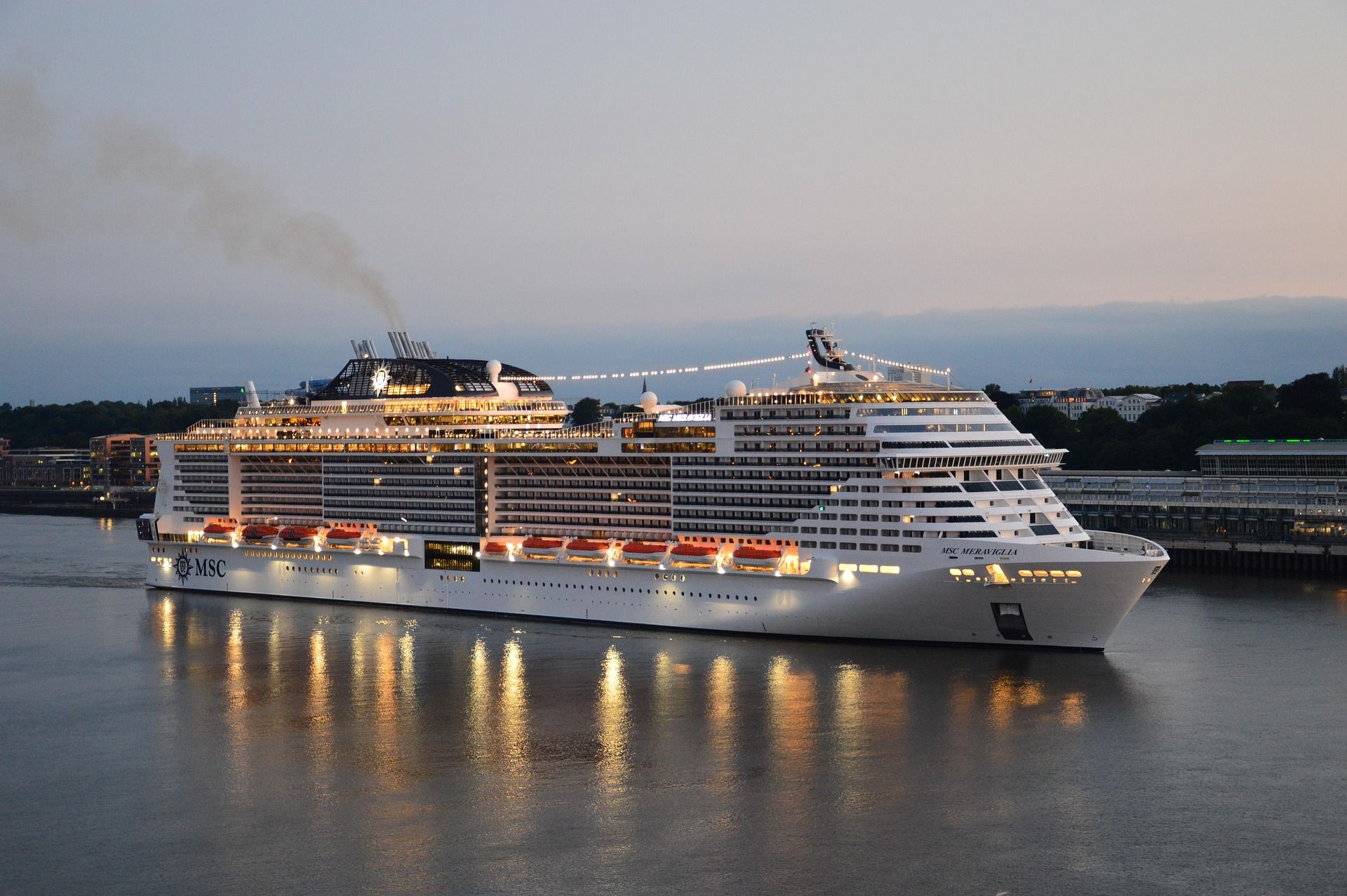 A large, brightly lit cruise ship sailing on calm water at dusk, with multiple decks, glowing window
