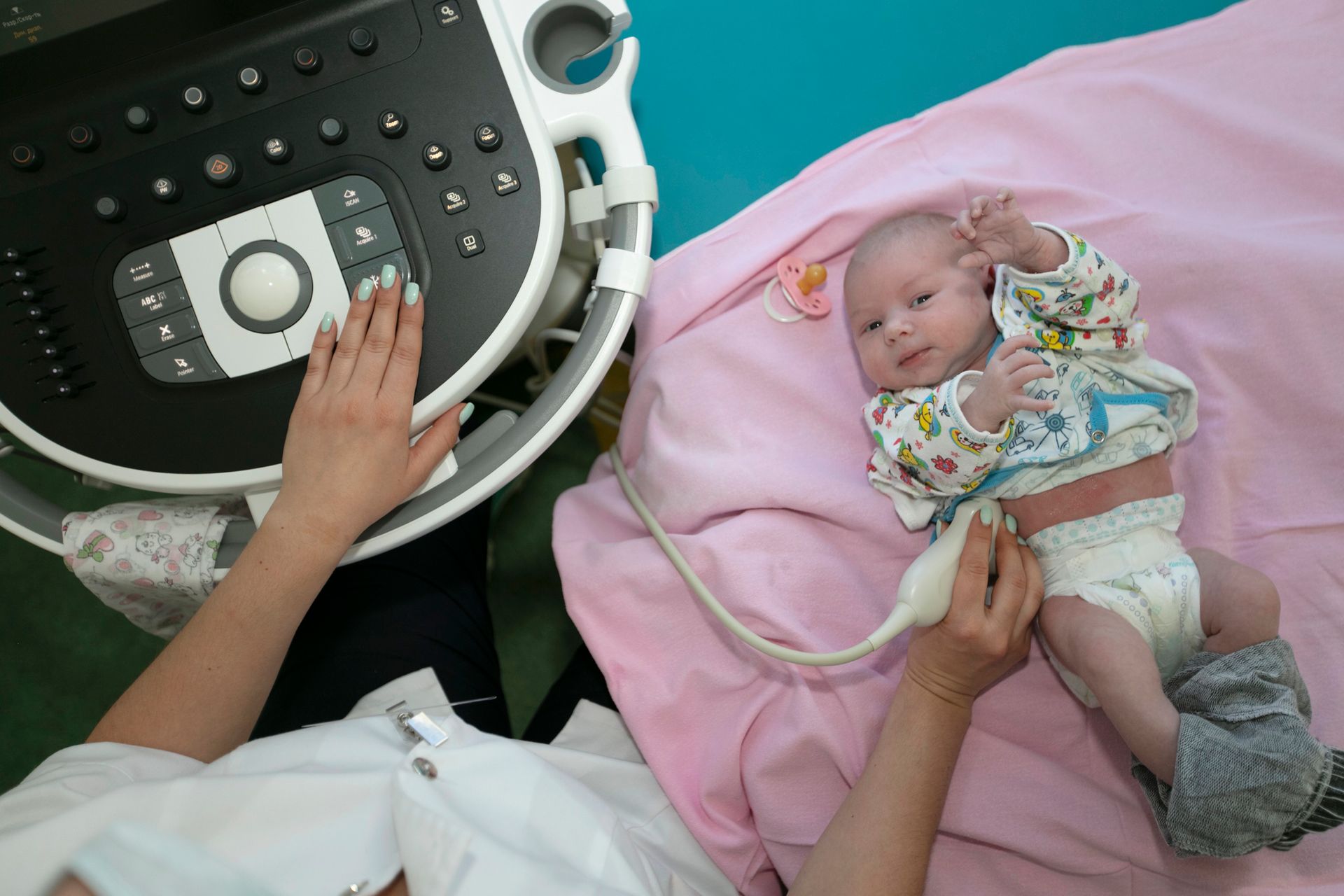 Mano di un adulto accanto a un neonato sdraiato su una coperta rosa, vicino a un monitor medico o a un ecografo nero