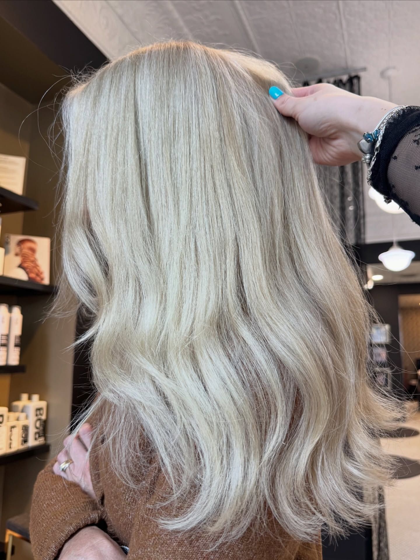 Blonde wavy shoulder-length hair being held up by a hand with blue nails in a salon setting