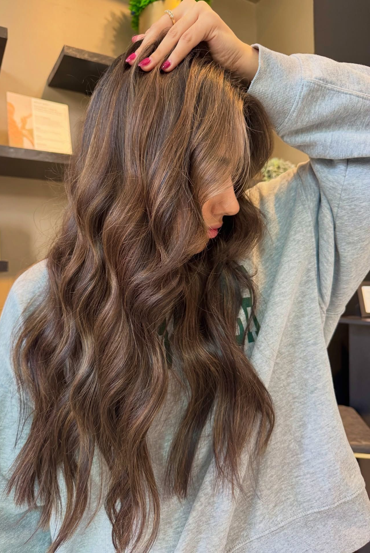 Woman with long wavy brown hair posing in a salon, wearing a gray sweater.