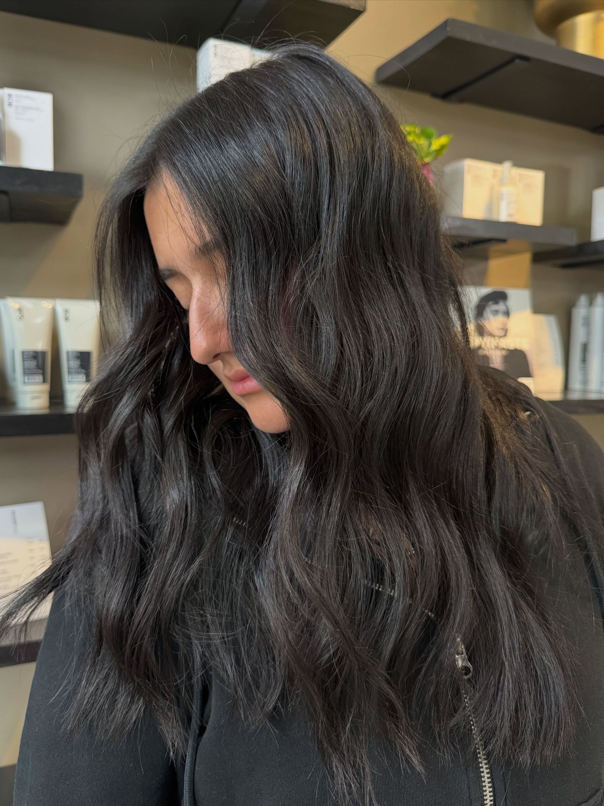 Woman with long dark wavy hair looking down in a salon, with product shelves in the background