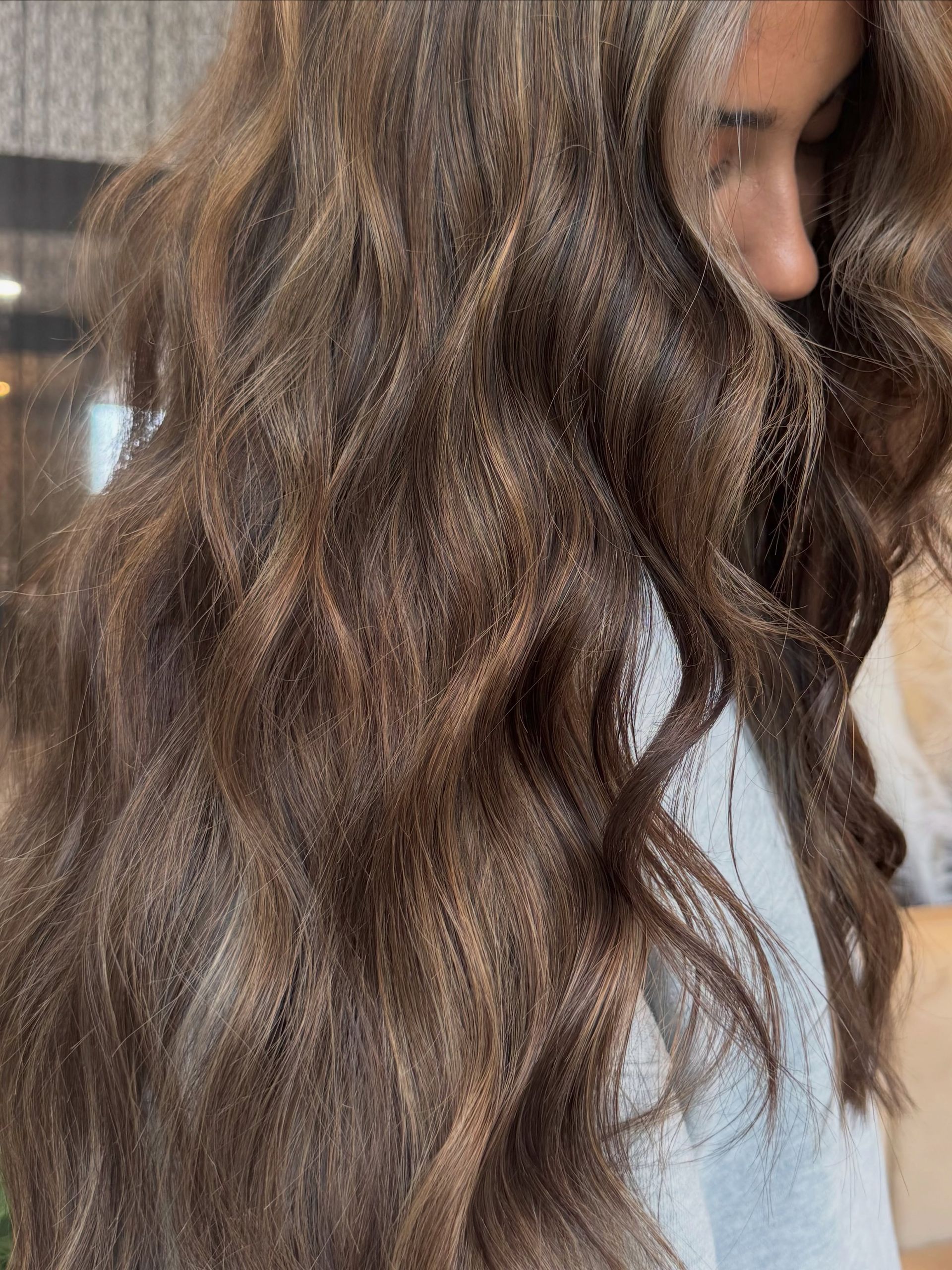 Side profile of long wavy brown hair with subtle highlights, flowing over a white shirt