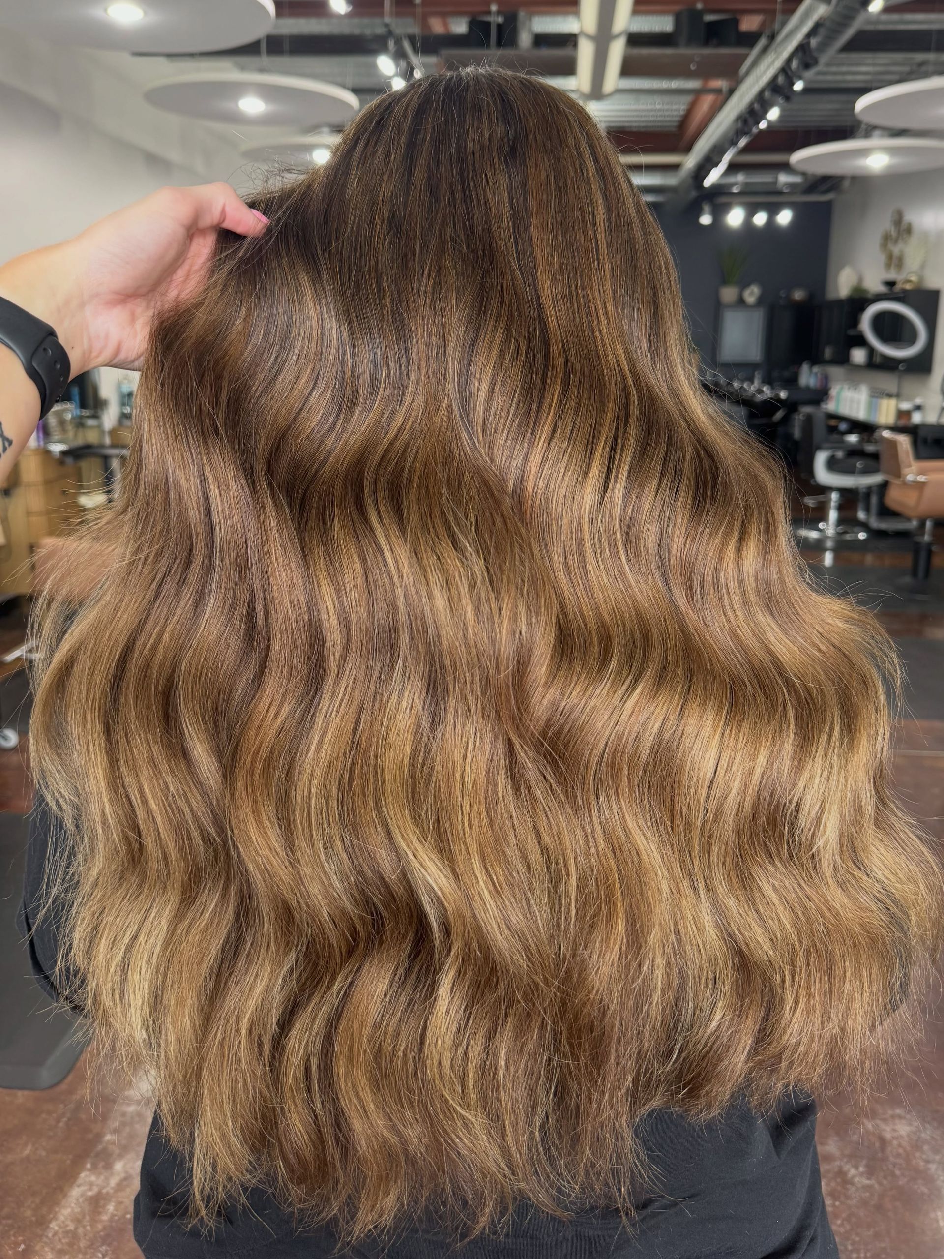 Long wavy brown hair viewed from behind in a salon chair, with a hand lifting the hair