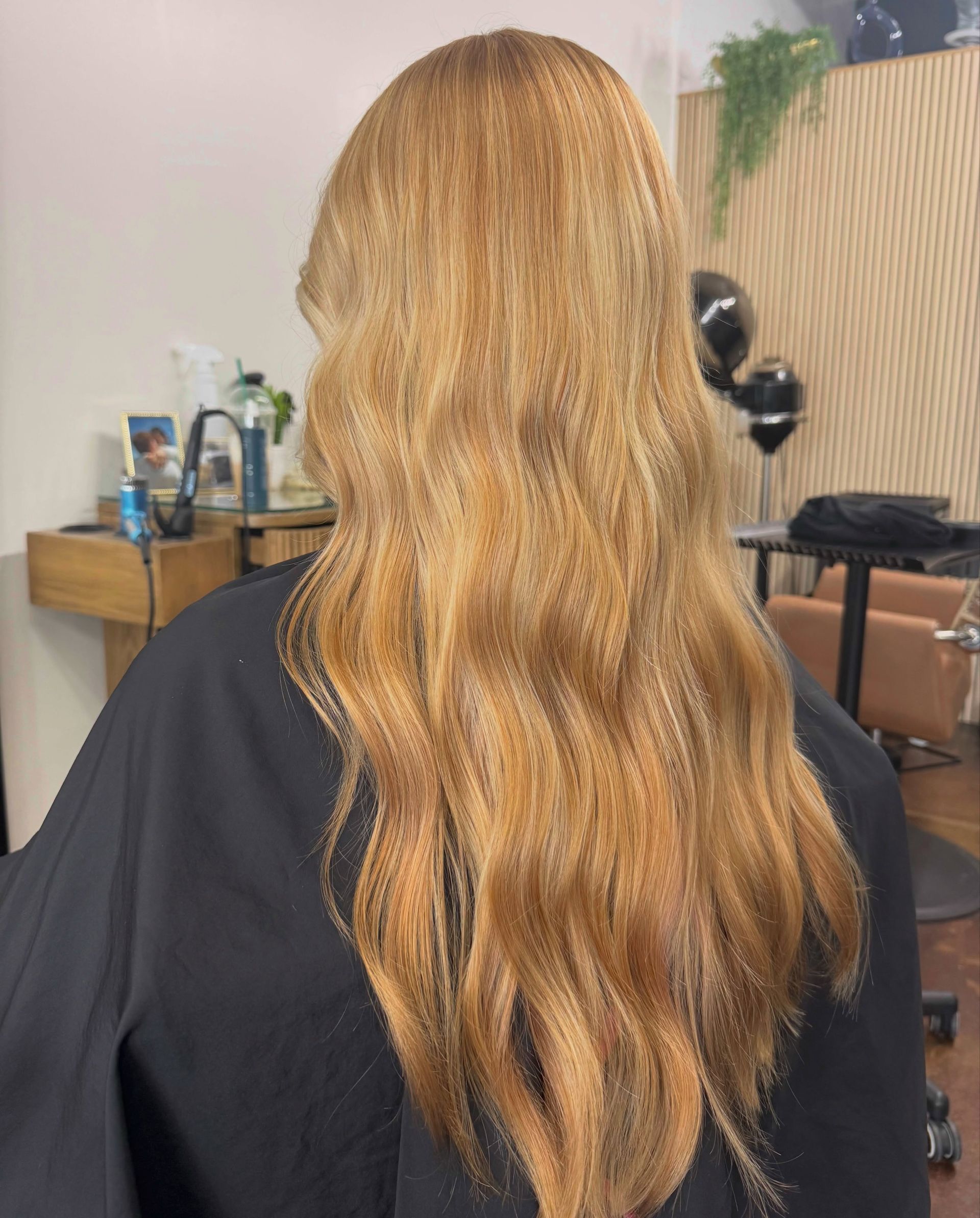 Long wavy blonde hair viewed from behind in a salon chair, with mirrors and styling tools in the background