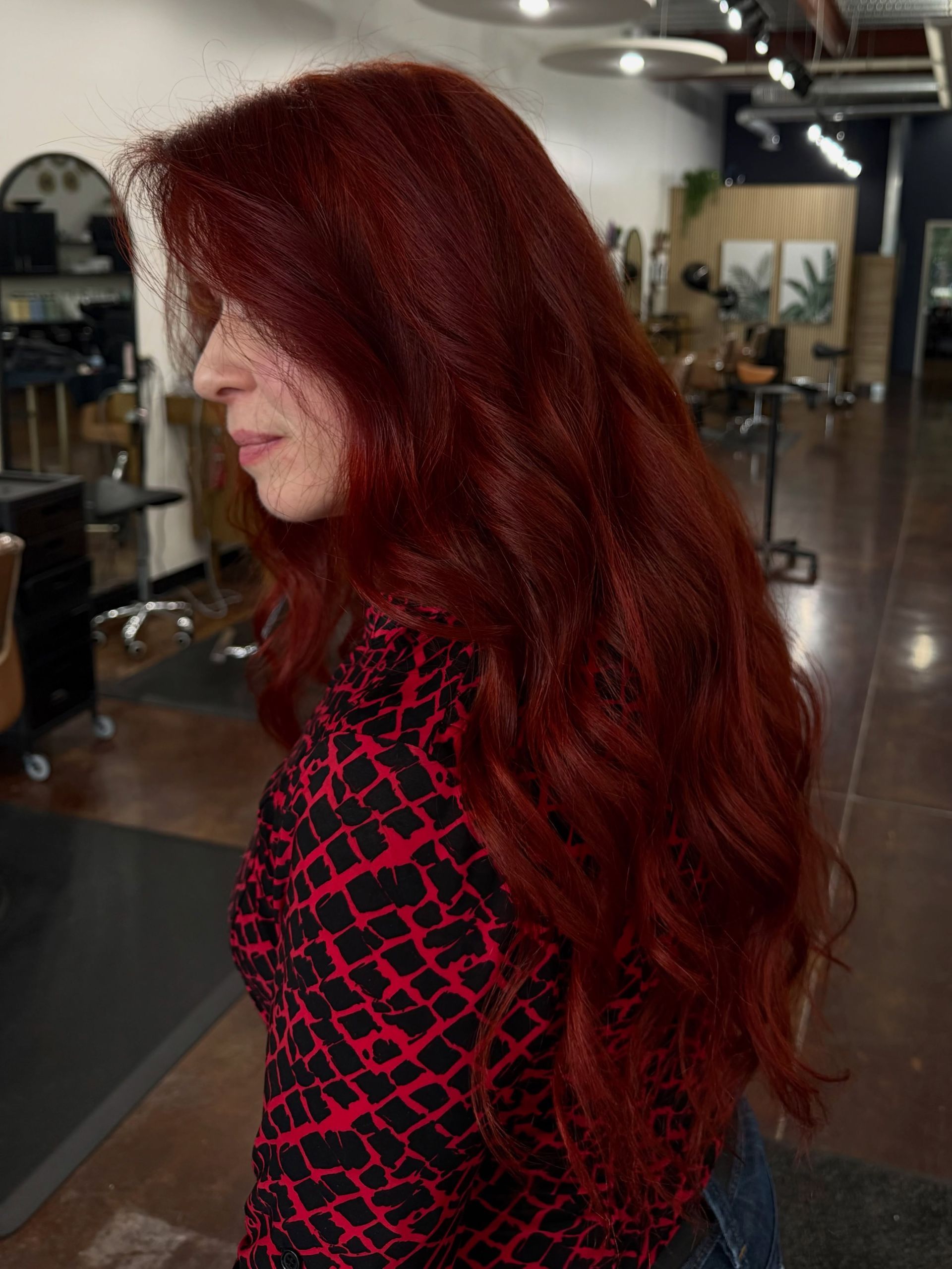 Woman with long red hair in a salon, wearing a black-and-pink patterned top.