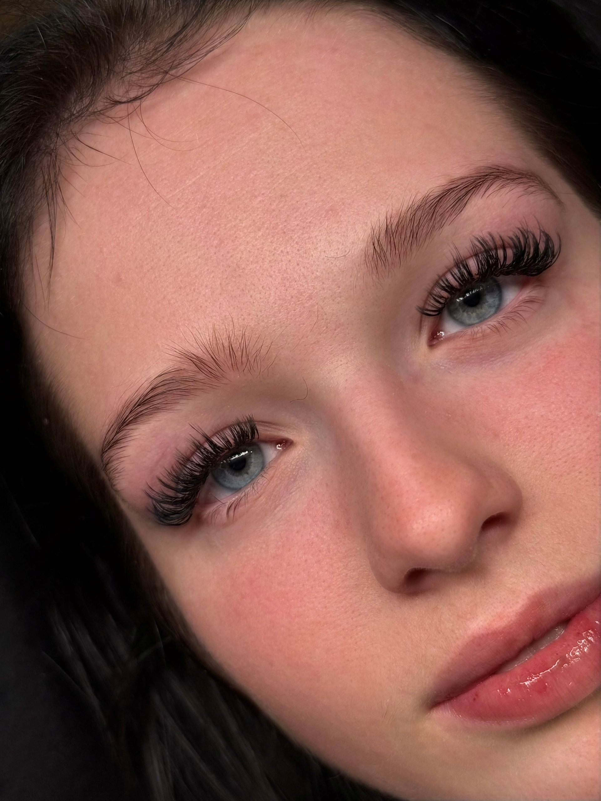 Close-up of a person with dark hair, soft makeup, and blue eyes, looking at the camera with a neutral expression