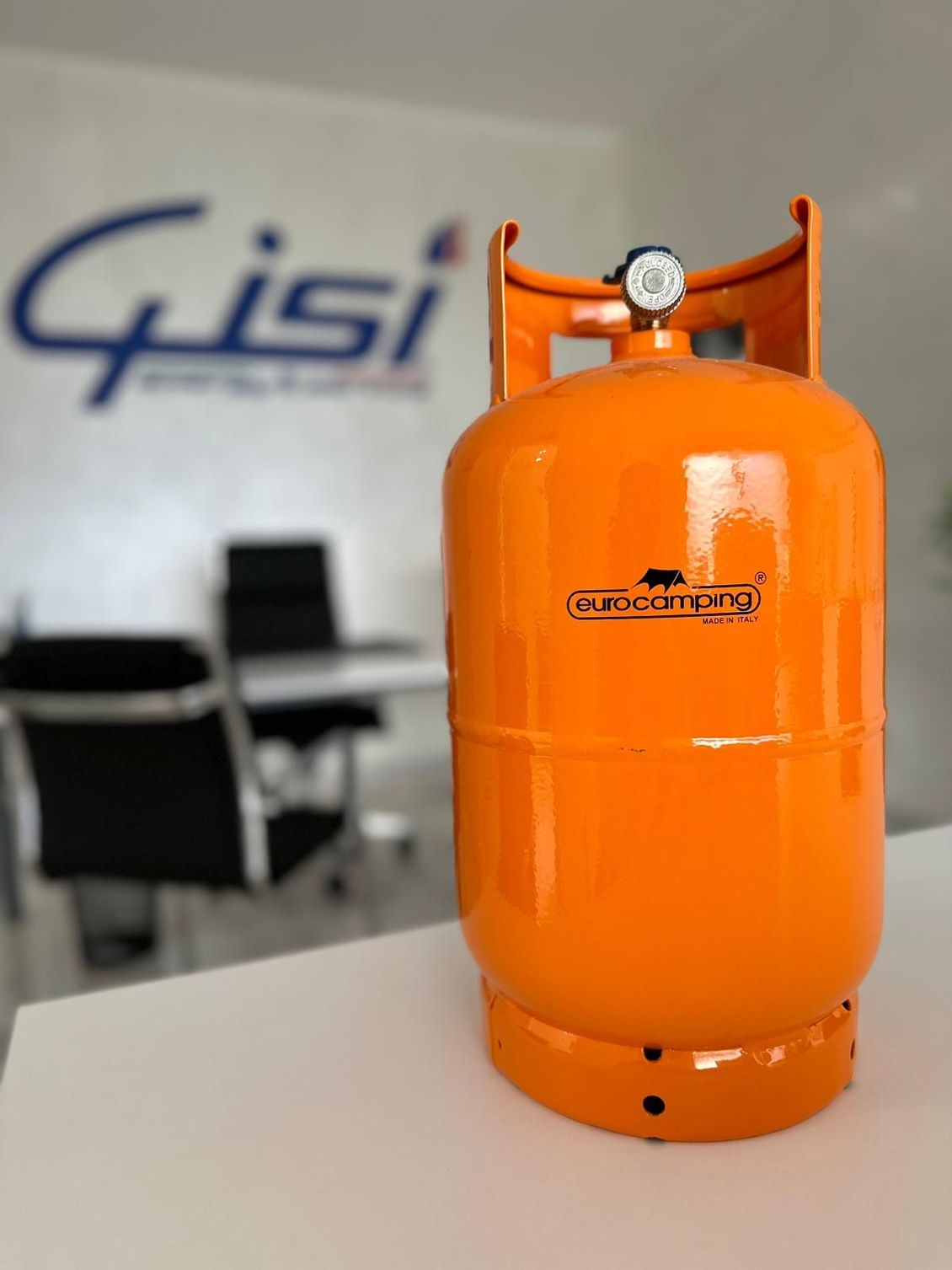 In un ufficio, una bombola di gas arancione è appoggiata su una superficie bianca, mentre sullo sfondo sfocato della parete è presente un logo.