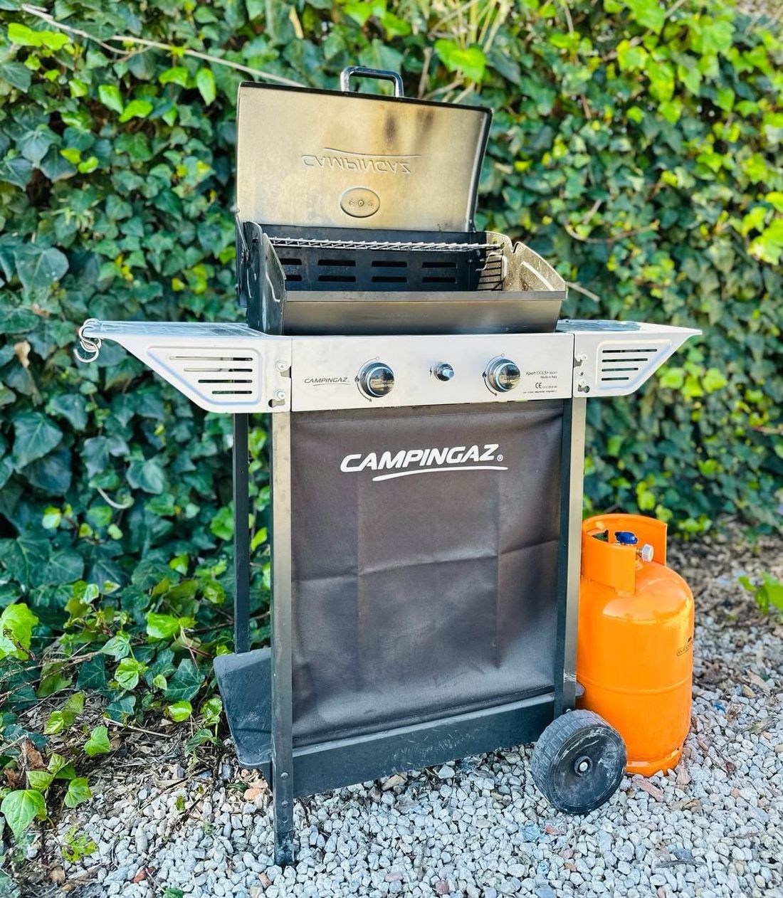 Un barbecue a gas Campingaz a due bruciatori con coperchio aperto, posizionato su un letto di ghiaia davanti a dell'edera, con una bombola di gas arancione nelle vicinanze.