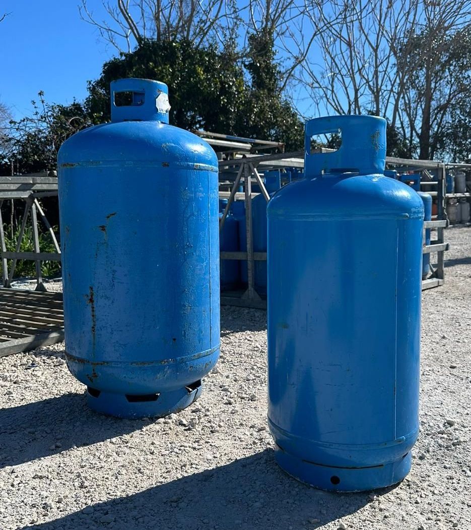 Due bombole di gas in metallo blu, appoggiate su una superficie di ghiaia chiara all'aperto, sotto un cielo azzurro brillante.