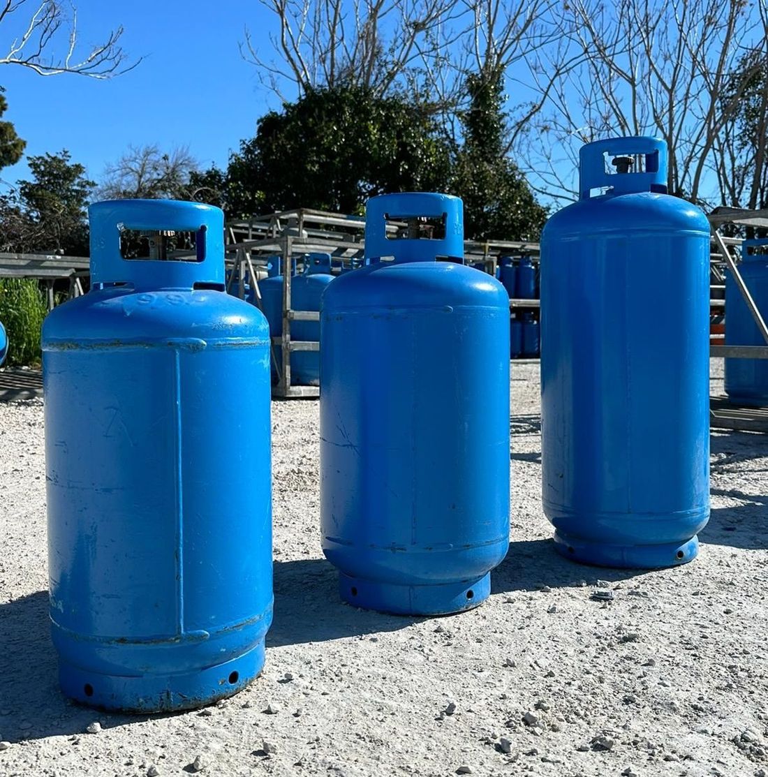 Tre bombole di gas blu si trovano all'aperto su un terreno ghiaioso sotto un cielo azzurro e limpido.