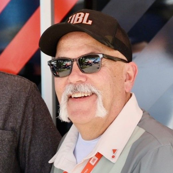 A smiling person wears an orange and black baseball cap and a textured red collared shirt, shown against a neutral wall.