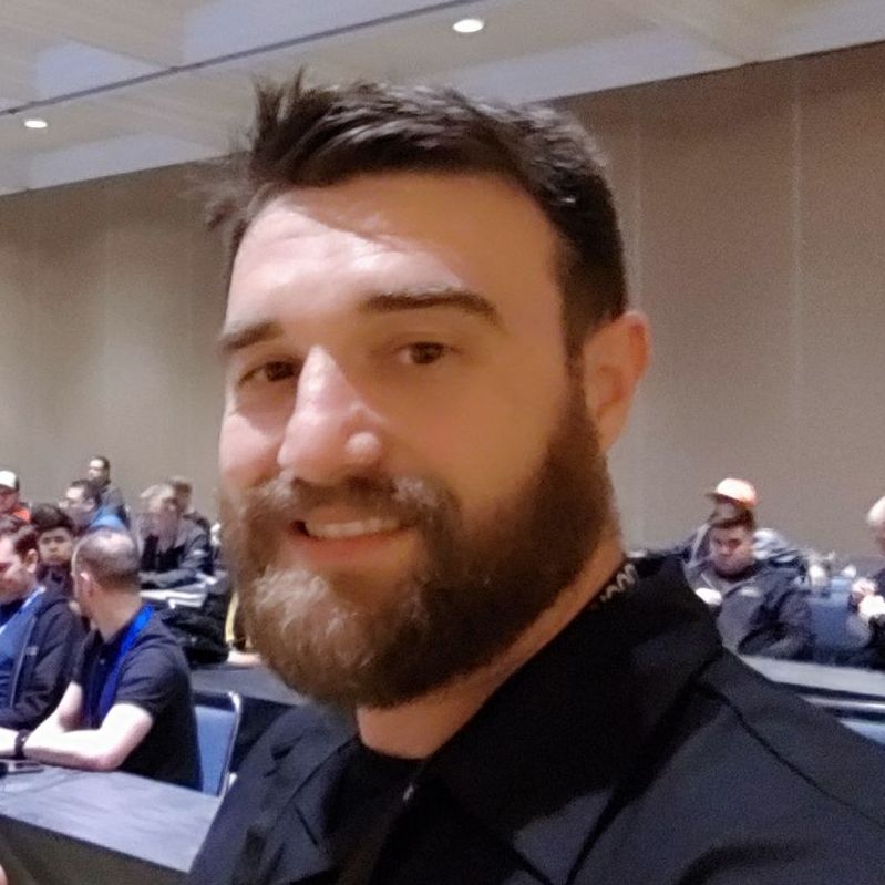 A bearded man smiles while sitting in a classroom or conference setting with other people in the background.