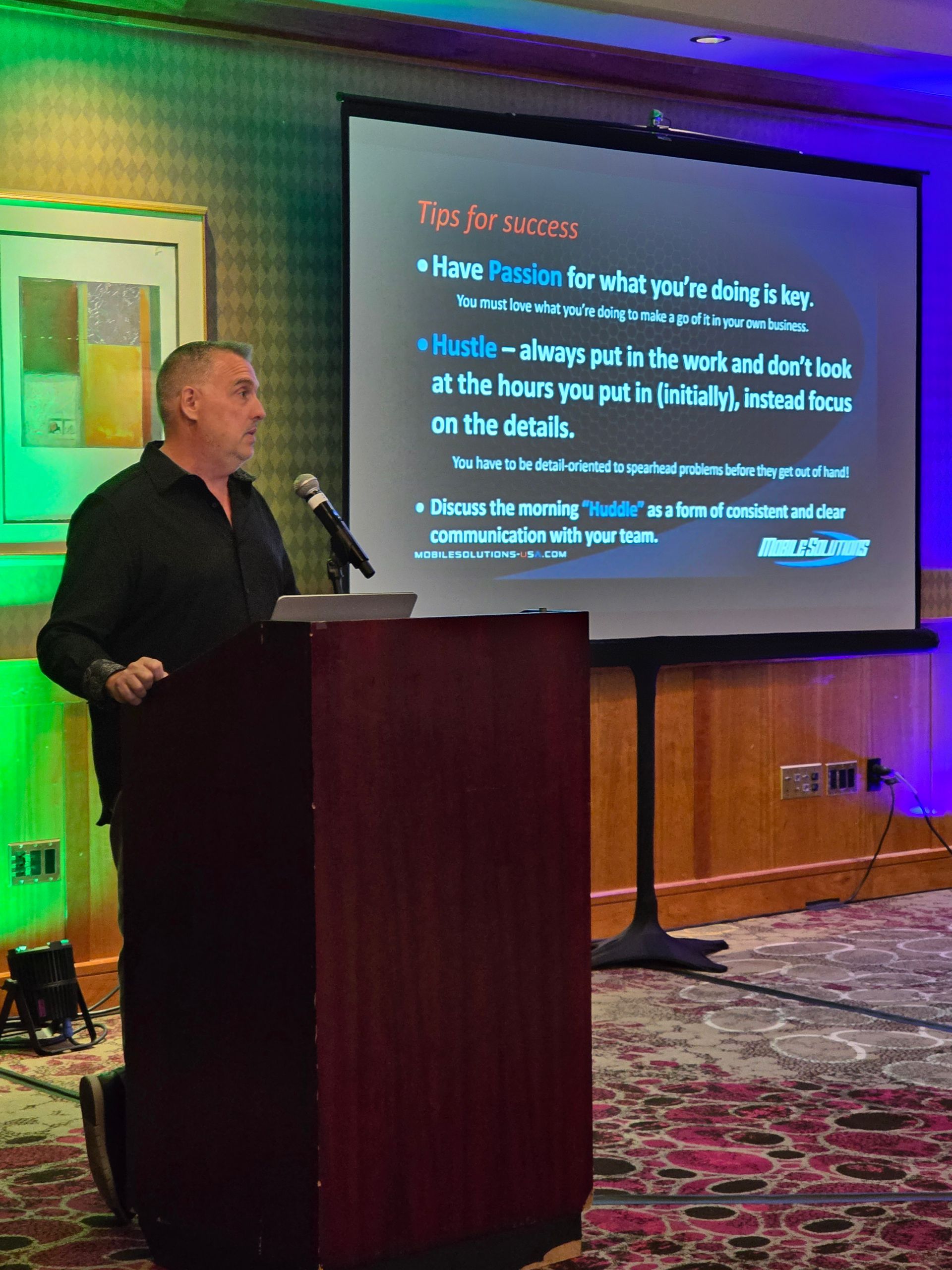 A man stands at a podium giving a presentation to an audience, with a screen behind him displaying tips for success.