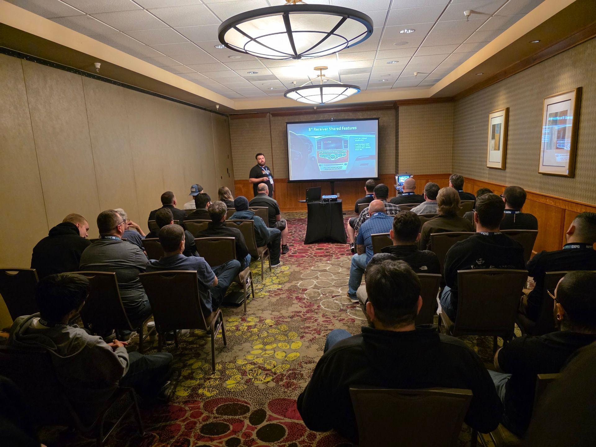A speaker presents to an audience seated in a conference room with a large screen and patterned carpeting.