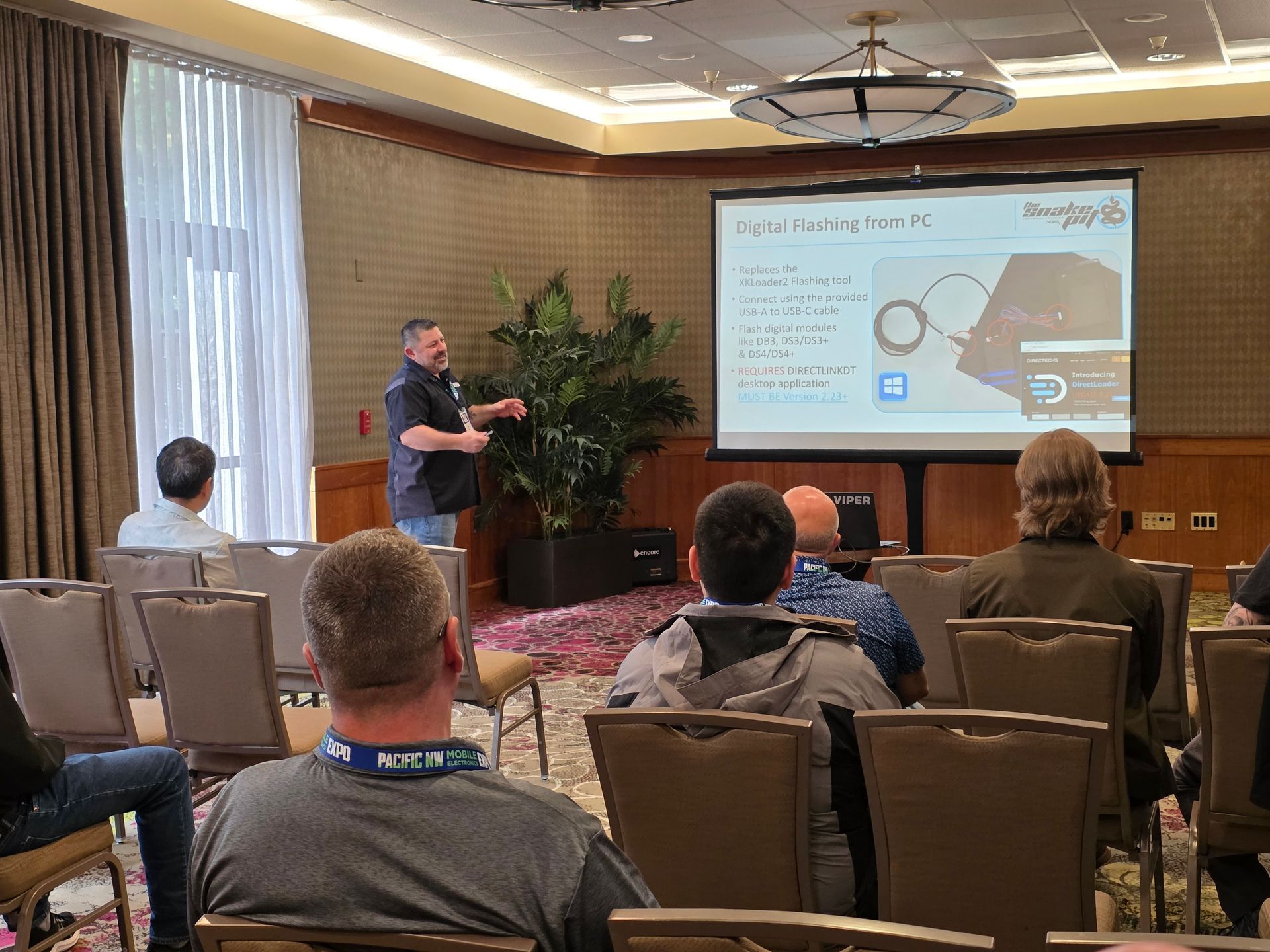 A presenter speaks to an audience in a conference room while gesturing toward a screen showing a technical diagram.