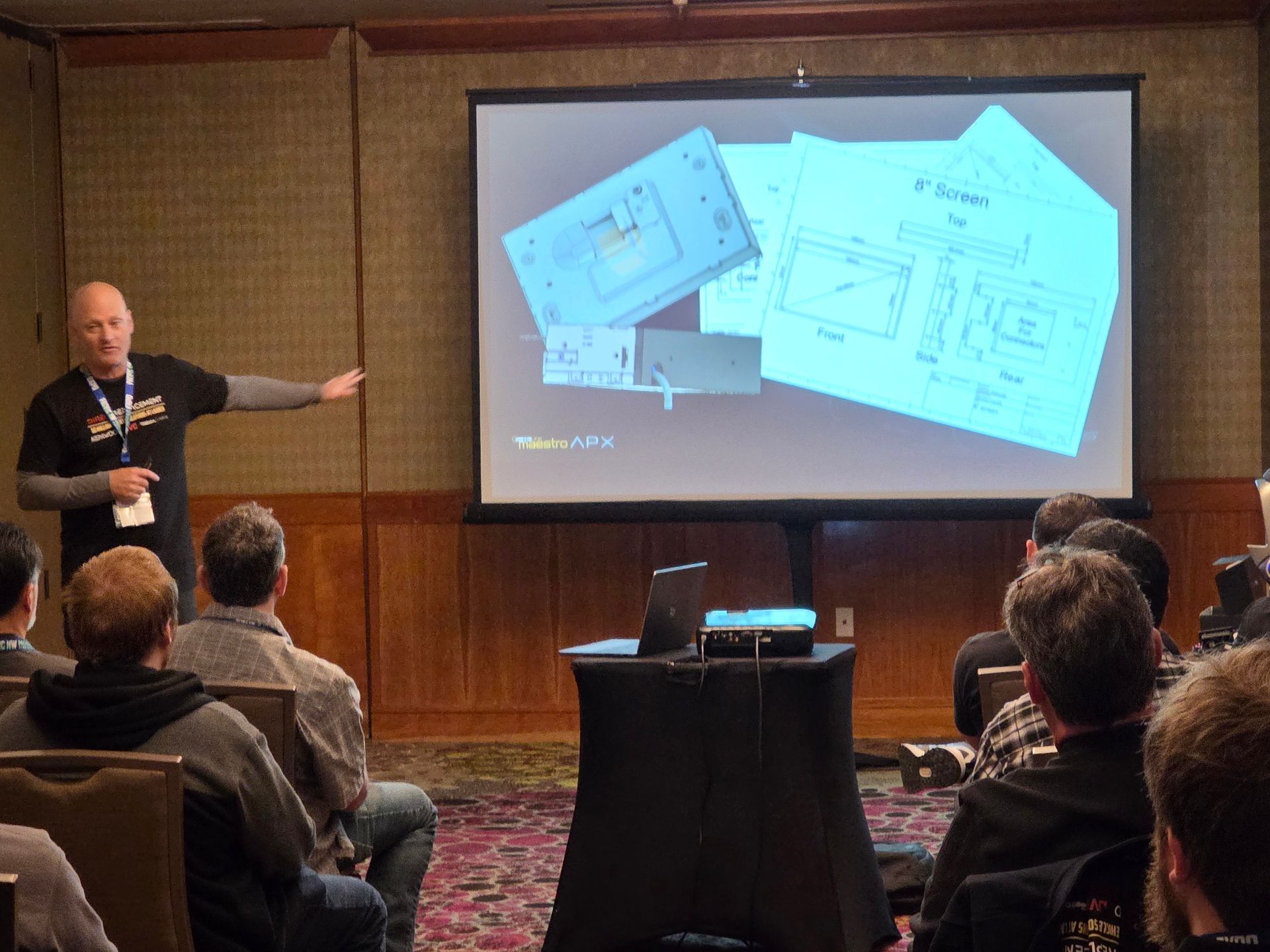 A presenter gestures toward a projected slide showing architectural floor plans to an audience in a conference room.