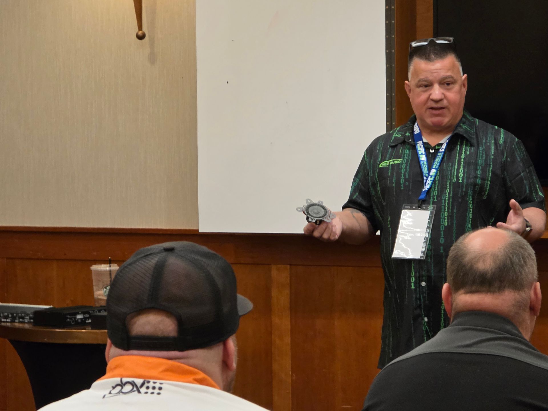 A person presenting to a group in a room, holding a small object while wearing a patterned shirt and lanyard.
