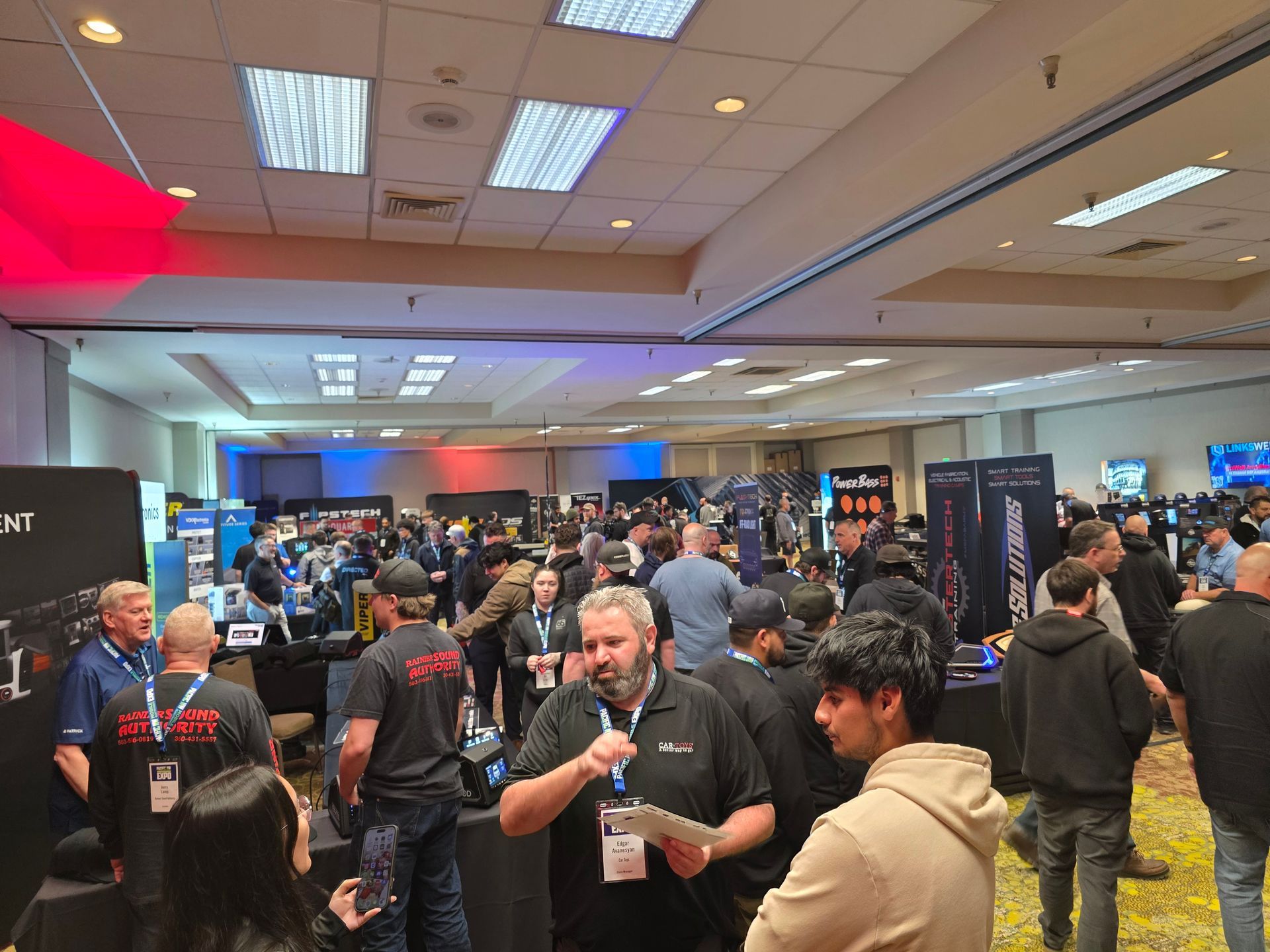 A crowded convention hall filled with people walking between vendor booths and talking at an indoor trade show.
