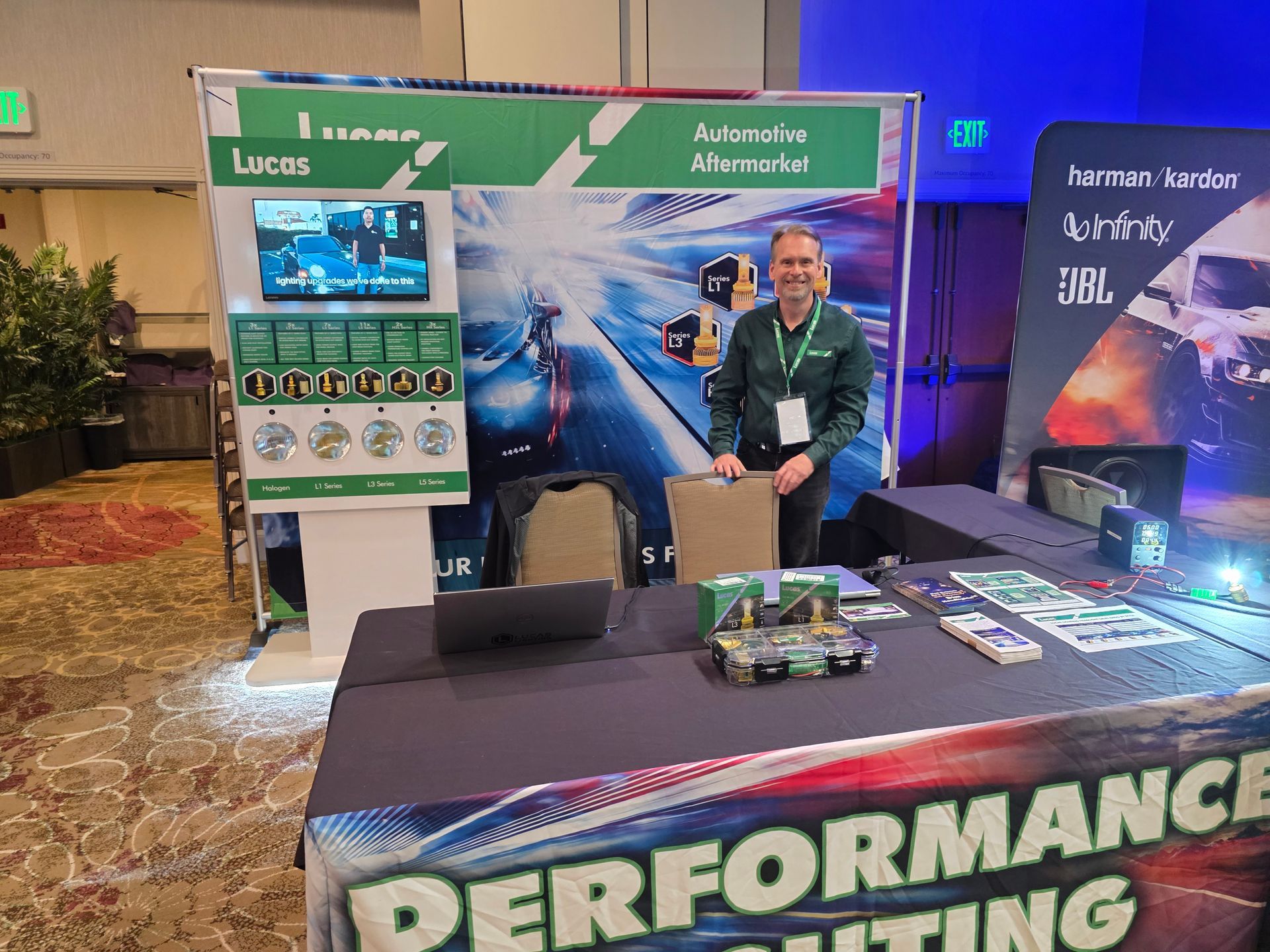A person stands at a trade show booth for Lucas Oil, featuring branded signage, a monitor, and a table with products.