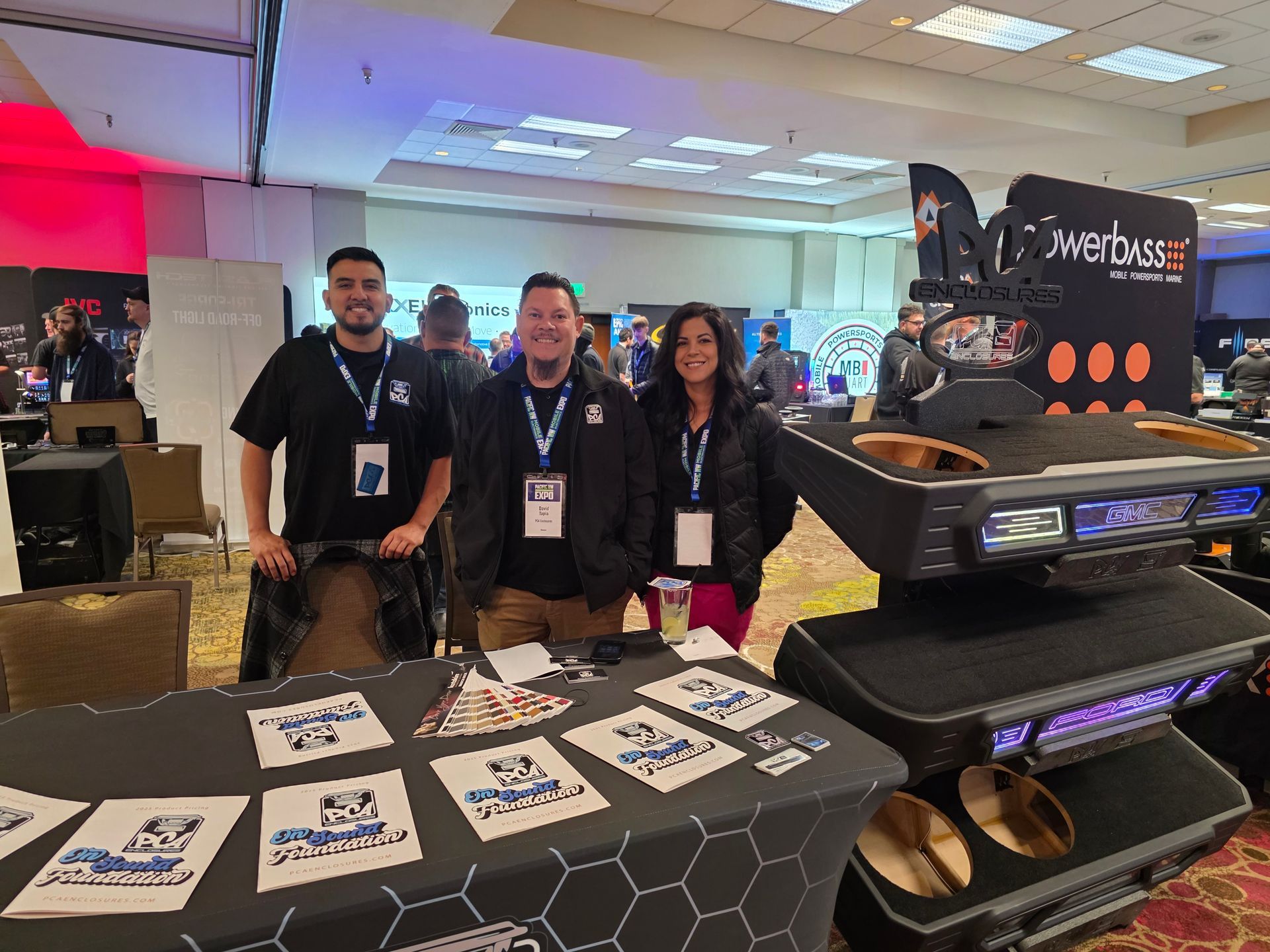 Three people stand behind a table with branding and display equipment at a trade show.