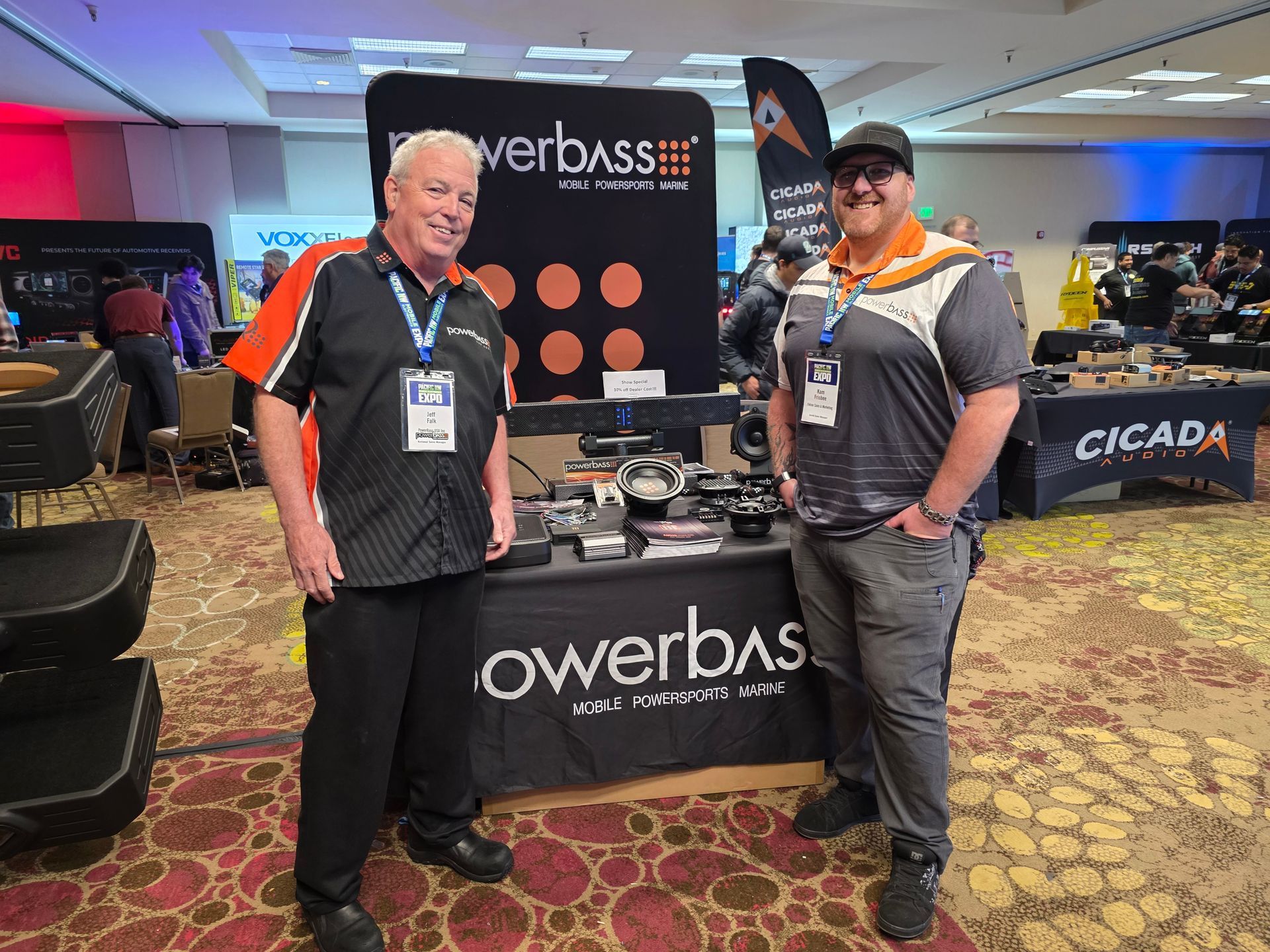 Two people stand smiling at a Powerbass booth at an indoor trade show with audio equipment on the table.