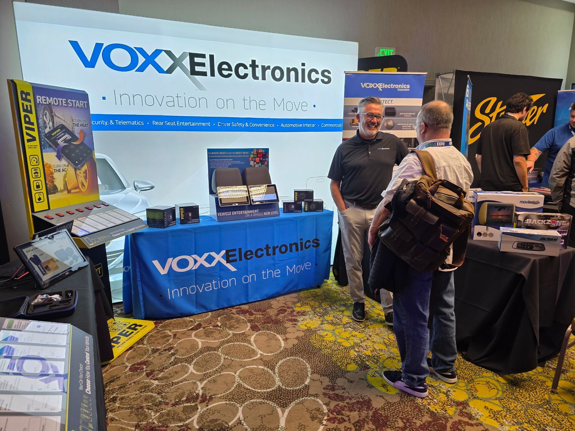Two people stand at a branded VOXX Electronics booth at a trade show, engaged in conversation near a display table.