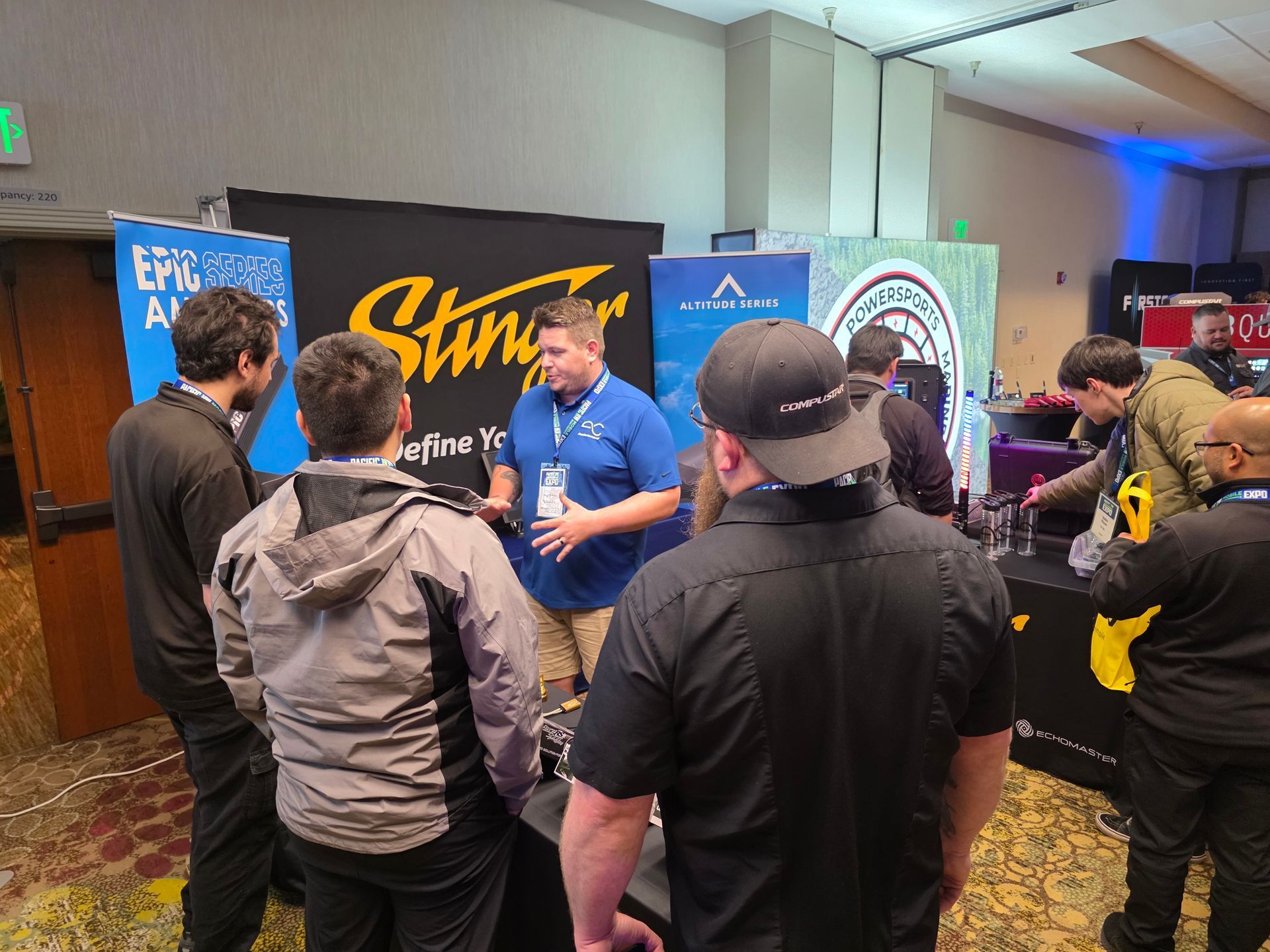A group of people gathers around a man speaking at a Stingray brand trade show booth in a conference room.