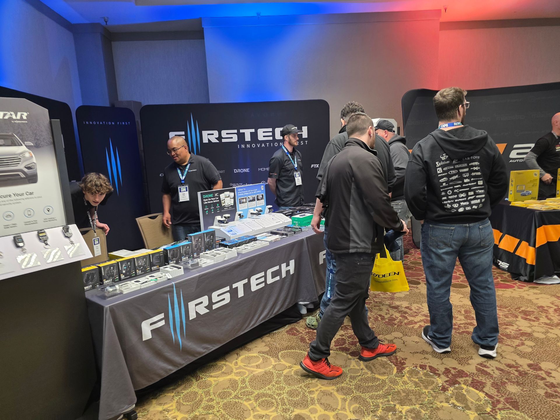 A Firstech booth at a trade show with staff and attendees interacting over a table display.