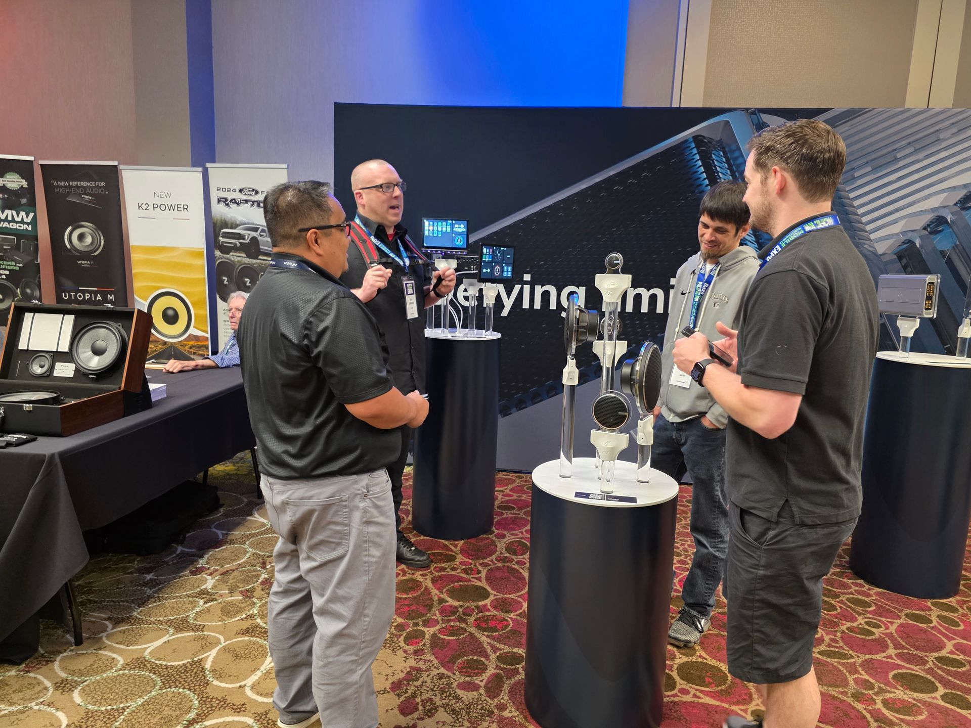 Four people stand around a display of audio equipment at a trade show exhibit, engaged in a professional conversation.