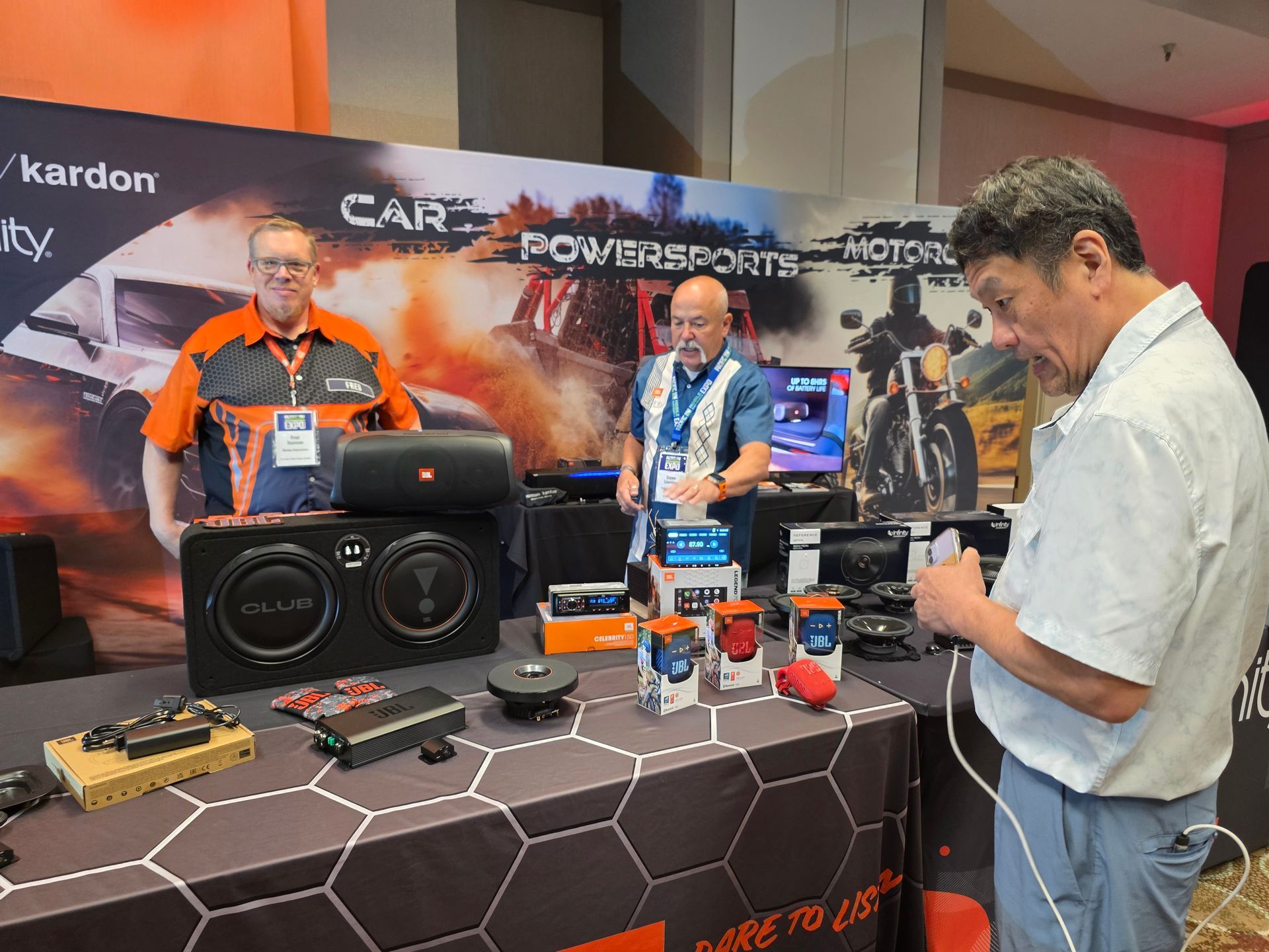 Three people at a trade show booth display JBL audio equipment, including speakers and amplifiers on a branded table.